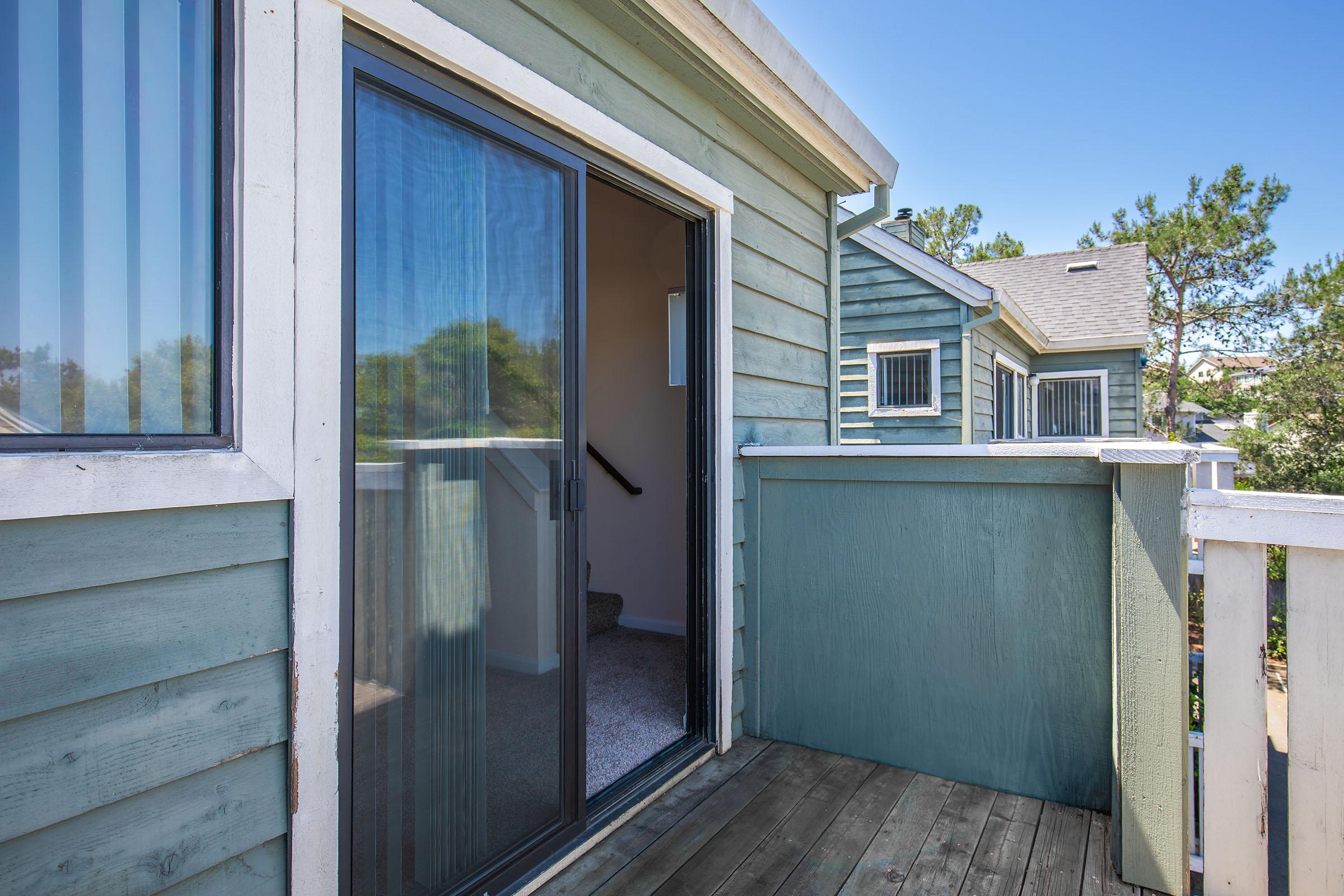 A view from a balcony with a sliding glass door, featuring wooden decking and a teal-colored railing. In the background, a glimpse of nearby houses with green and blue siding is visible, surrounded by trees under a clear blue sky.