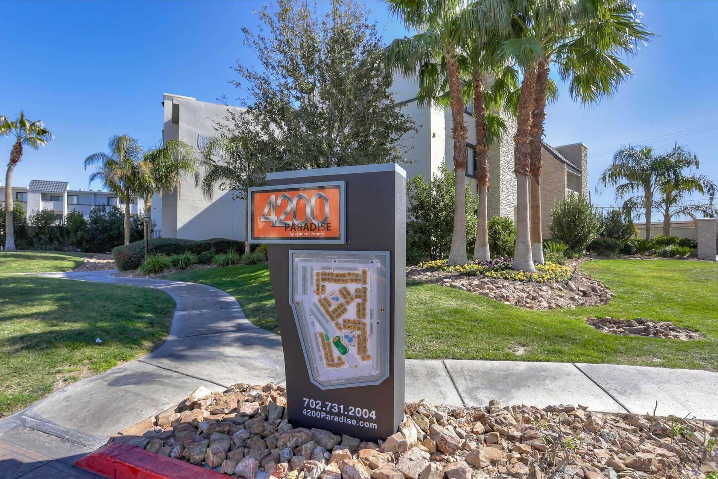 Sign for "4200 Paradise" featuring a map of the property layout, surrounded by landscaped greenery and palm trees. The sign includes contact information: phone number 702.731.2004. The area appears well-maintained with a landscaped pathway leading to the sign.