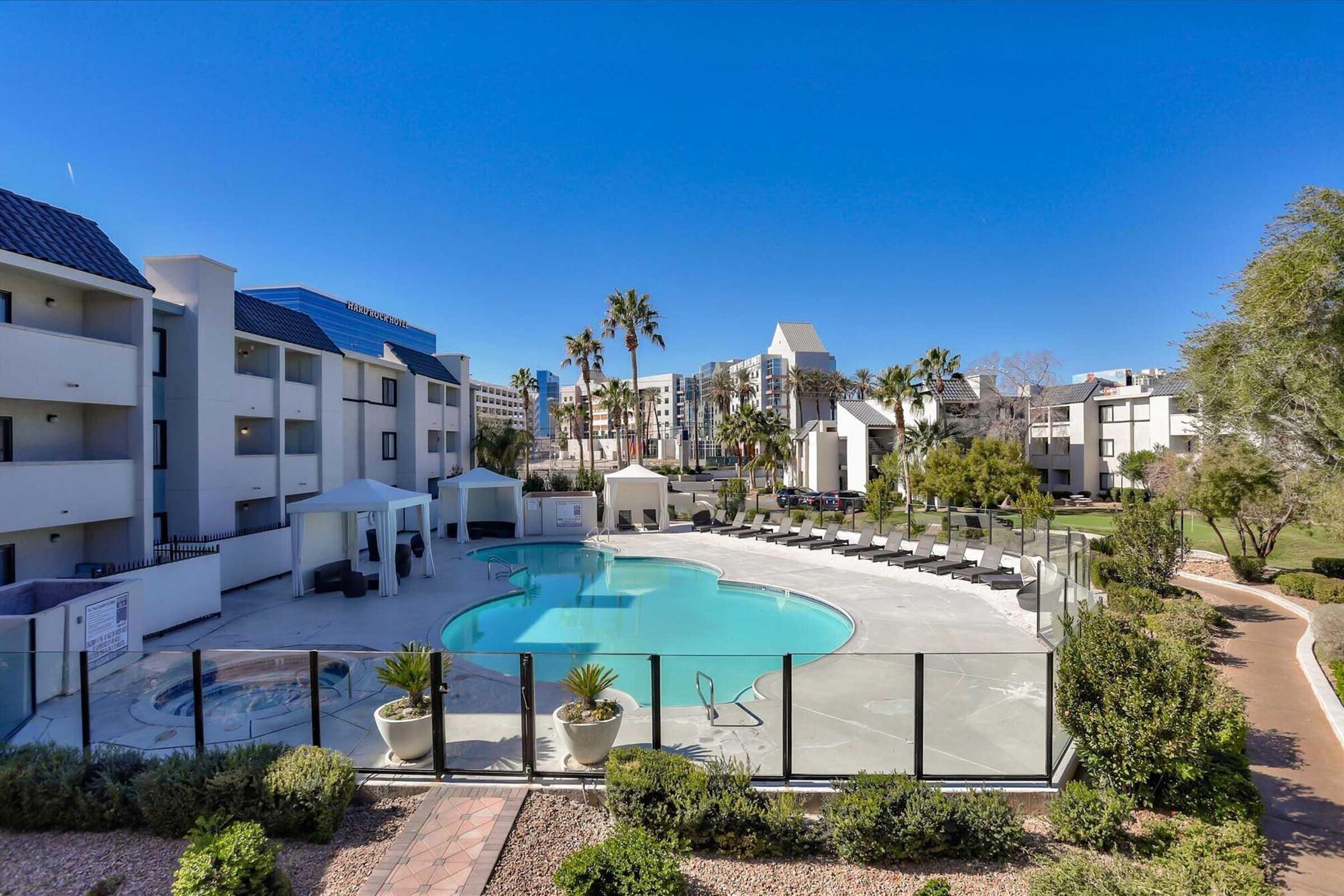 A clear blue sky above a well-maintained swimming pool surrounded by lounge chairs and palm trees. The pool area features cabanas and is enclosed by a glass fence. Nearby, modern buildings can be seen, creating a modern resort-like atmosphere.