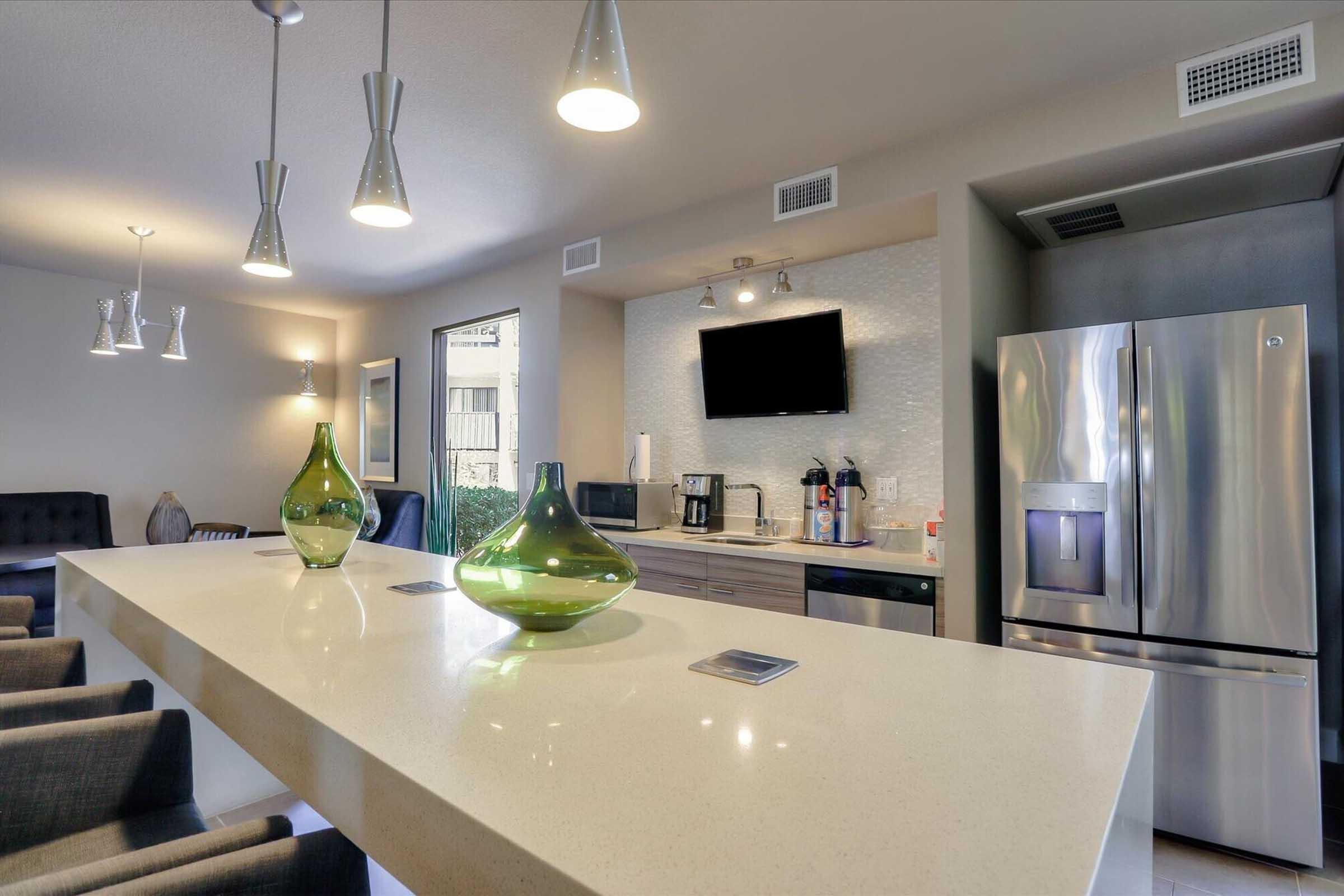 A modern kitchen with a long countertop featuring green decorative vases, pendant lighting, and a sleek silver refrigerator. The space includes a coffee maker and other appliances, with a small television mounted on the wall. The décor is contemporary and inviting, with natural light coming from a window.