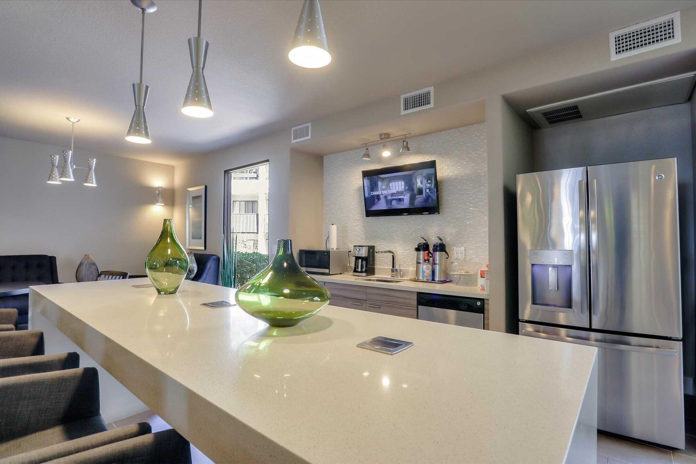 A modern kitchen featuring a large white countertop with two green decorative vases. In the background, there is a sleek stainless steel refrigerator and a TV mounted on the wall. The space is well-lit with pendant lights and includes seating with gray chairs. A window offers a glimpse of greenery outside.