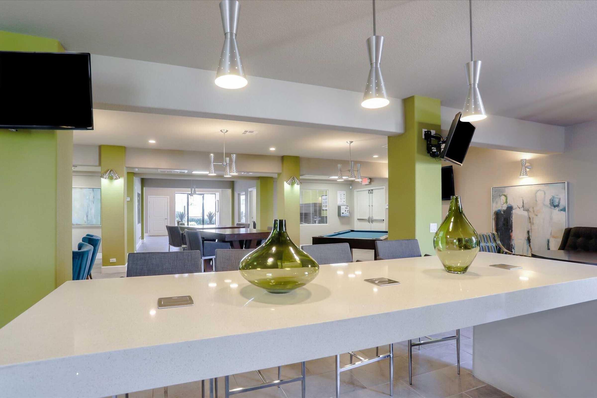 A modern lounge area featuring a sleek bar with three decorative vases, pendant lighting, and a flat-screen TV. The space includes seating arrangements and a pool table in the background, with bright green accents on the walls, creating a stylish and inviting atmosphere.