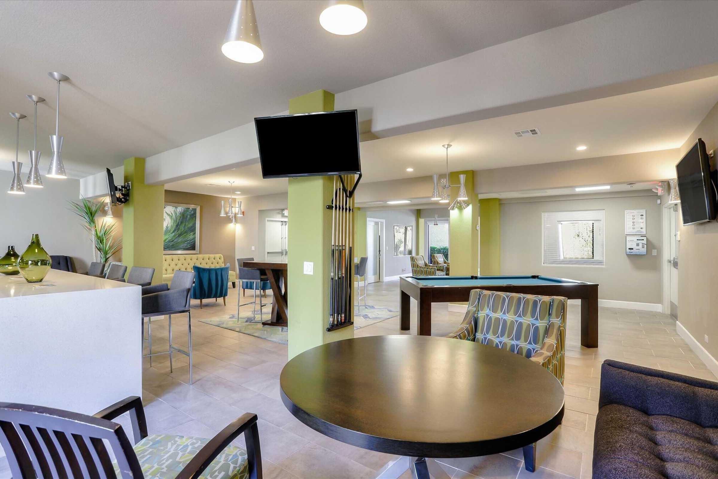 A modern common area featuring a pool table, tables, and comfortable seating. There are stylish light fixtures and televisions mounted on the walls. The space has a fresh, contemporary design with green accents, large windows for natural light, and decorative plants, creating an inviting environment for socializing.