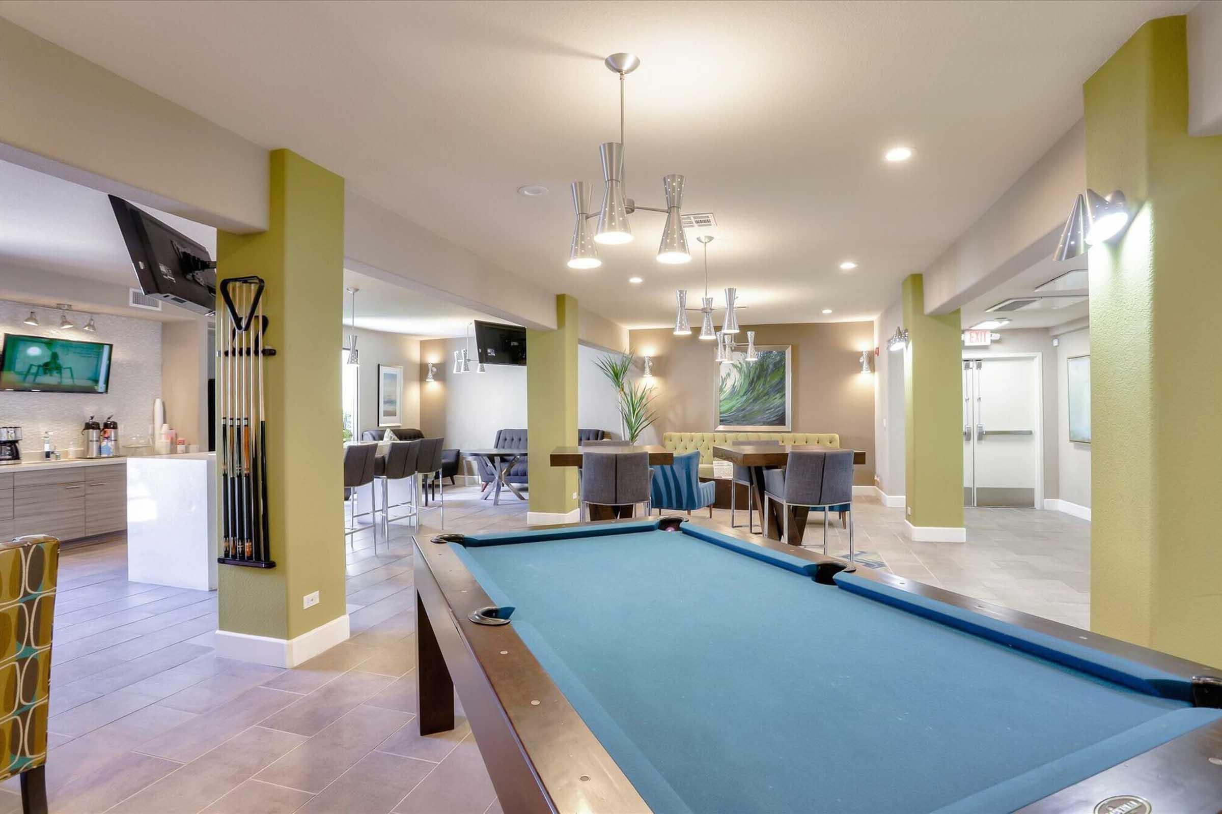 A spacious lounge area featuring a blue pool table, modern lighting, and seating arrangements. There are flat-screen TVs mounted on the walls, a bar area with high chairs, and large windows providing natural light. The décor includes green walls and contemporary artwork, creating an inviting atmosphere.