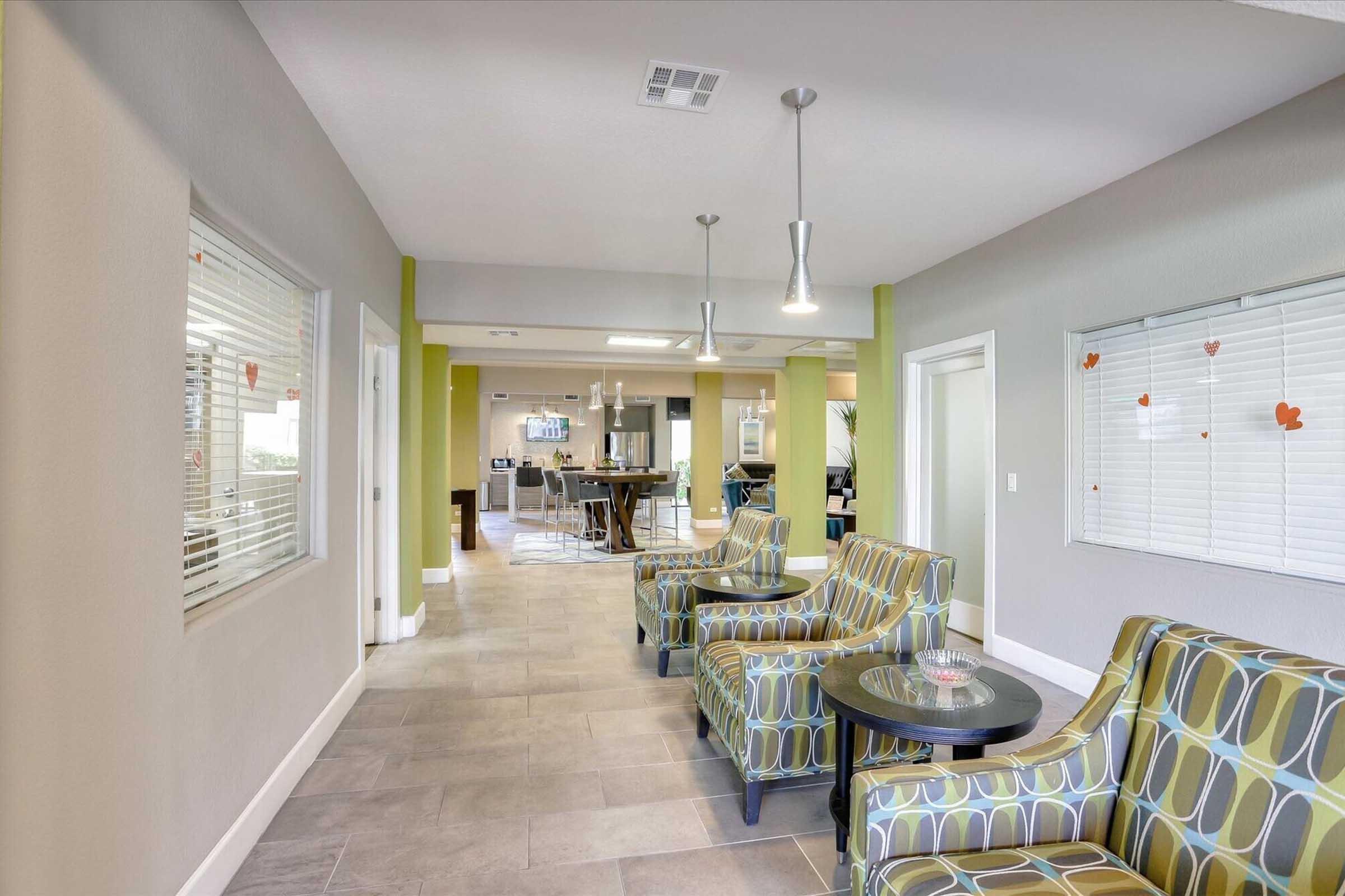 A modern lobby area featuring contemporary seating with vibrant patterned cushions. The space is well-lit with pendant lights and has light-colored walls. A large window lets in natural light, and colorful decorations hang on the walls. The atmosphere is inviting and stylish, suitable for socializing or relaxing.