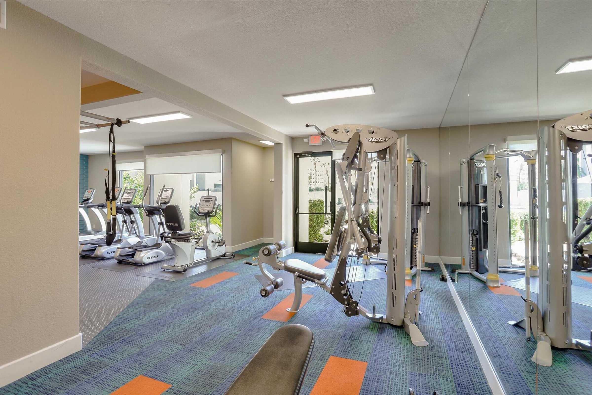 A modern gym interior featuring various exercise equipment, including weight machines and cardio machines. The space has bright lighting, mirrors on one wall, and a vibrant carpet design. Large windows provide natural light and a view of the outdoors.