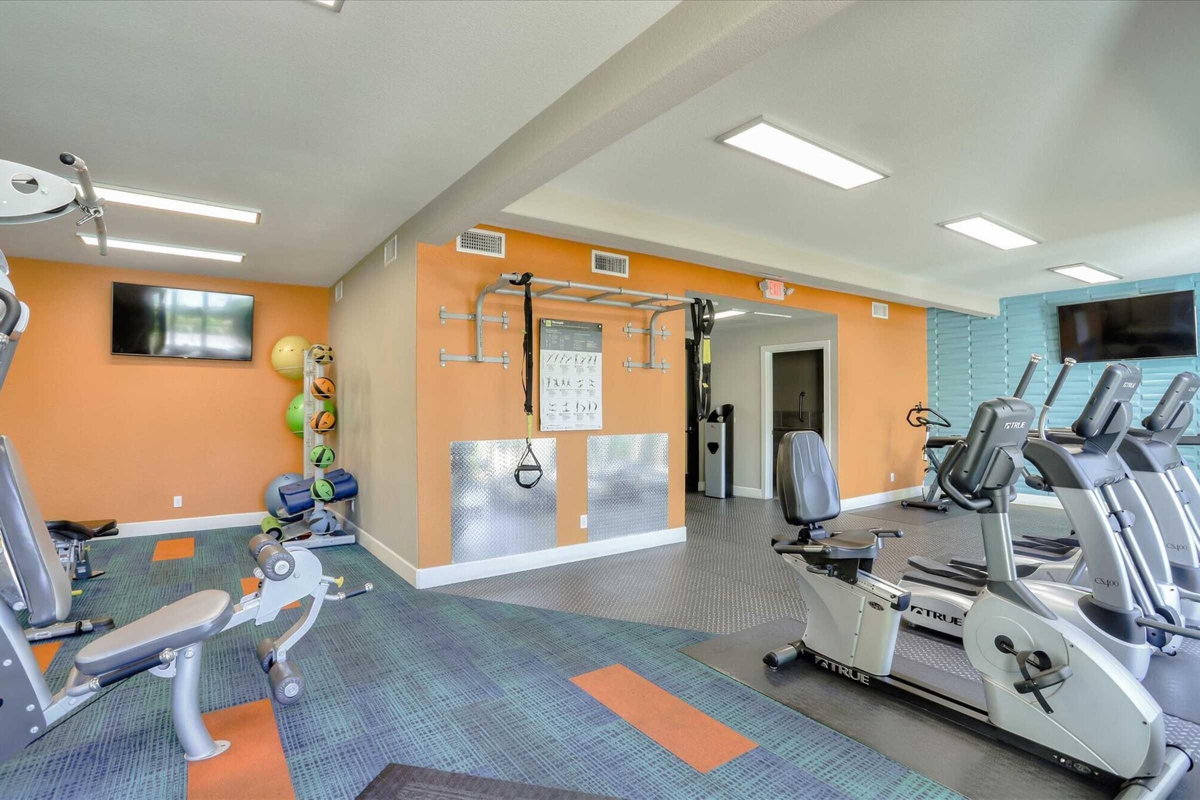Interior of a modern gym featuring various exercise equipment, including stationary bikes and rowing machines. The walls are painted orange and blue, with motivational posters and fitness accessories, such as stability balls, stored on shelves. Bright lighting enhances the open and inviting atmosphere.