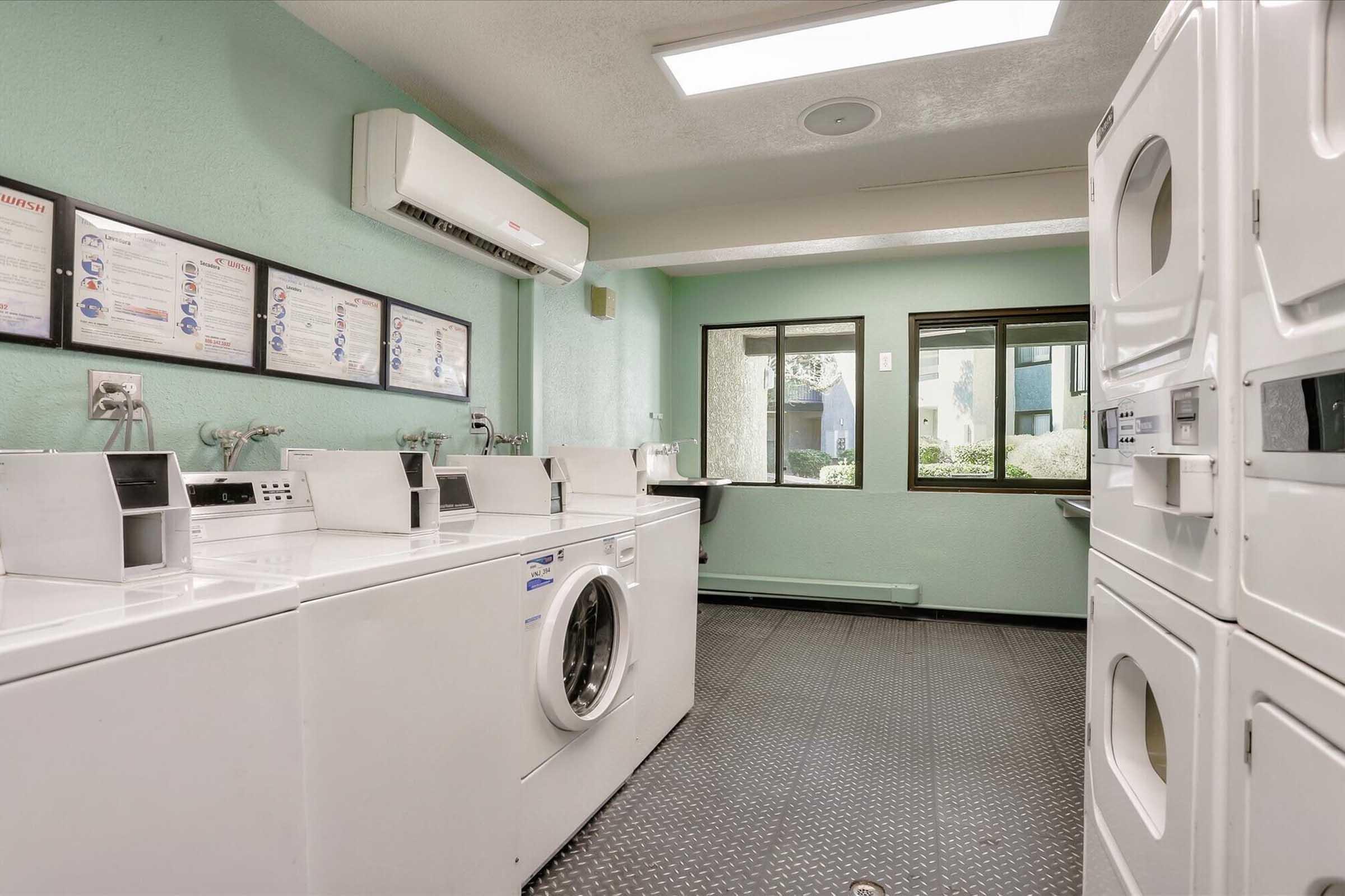 A bright laundry room featuring several white washing machines and dryers. There are sink areas along one wall, with a large window providing natural light and a view of greenery outside. Wall posters displaying laundry instructions and tips are visible. The floor is black with a textured pattern.