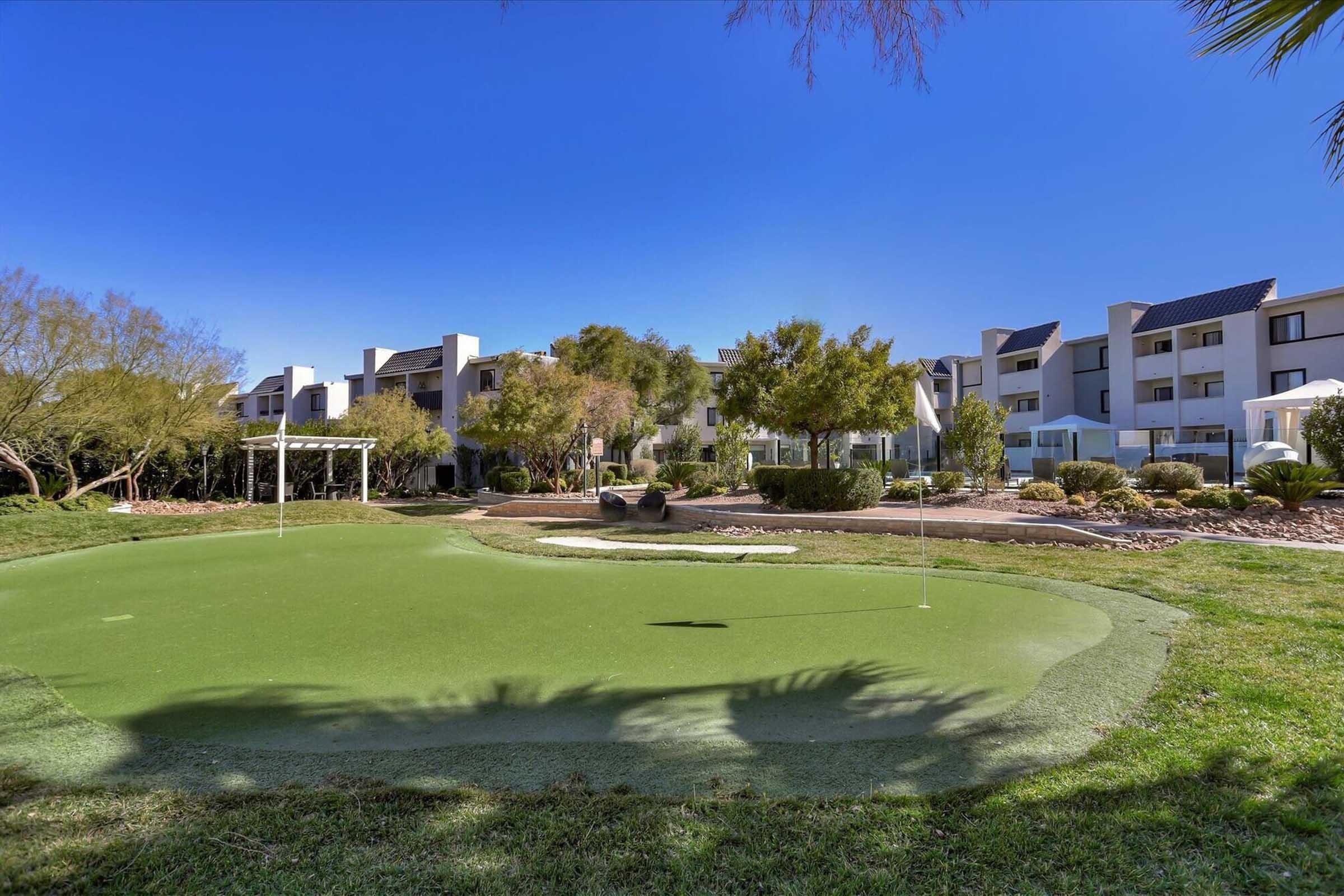 A well-maintained putting green surrounded by lush grass and landscaping, with modern-style buildings in the background under a clear blue sky. The scene reflects a serene and inviting atmosphere, ideal for relaxation and leisure activities.