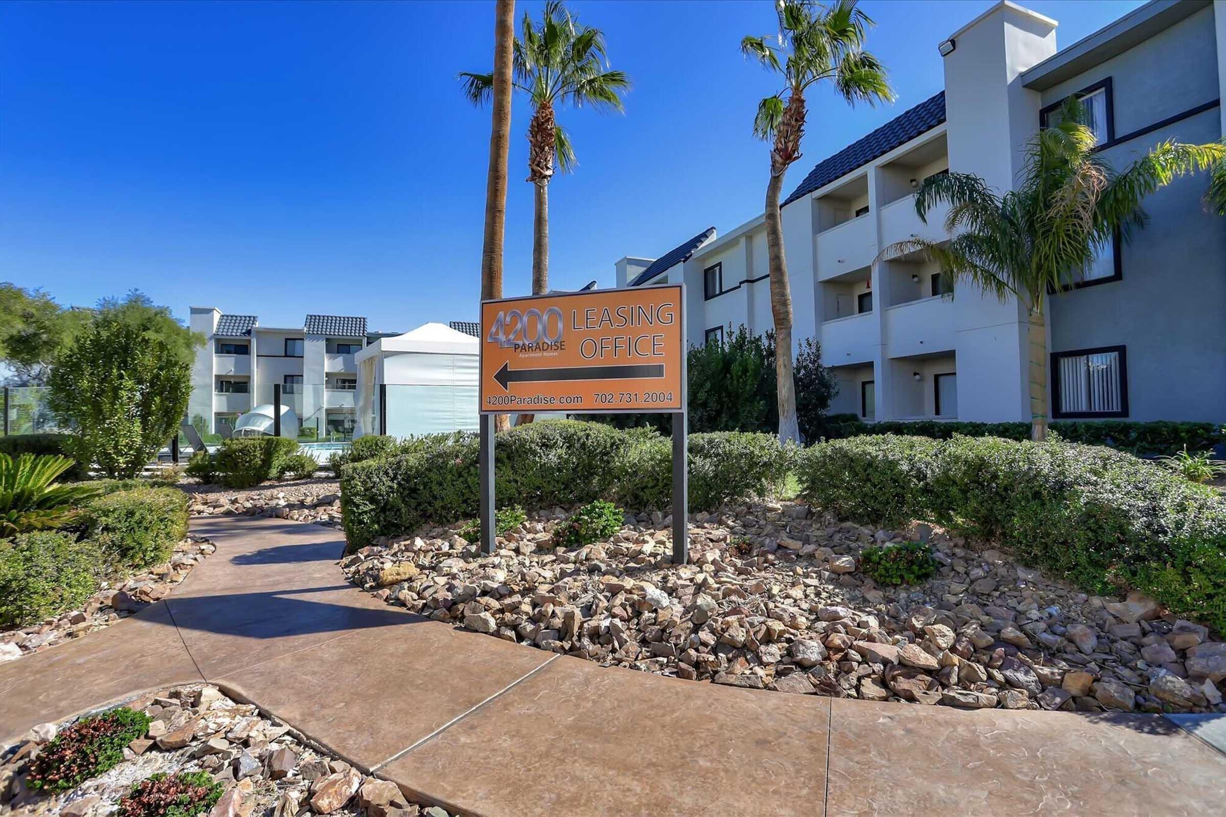 Leasing office entrance with a large sign displaying "Leasing Office." The pathway is lined with rocks and surrounded by neatly manicured shrubs and palm trees. In the background, there are modern apartment buildings under a clear blue sky.