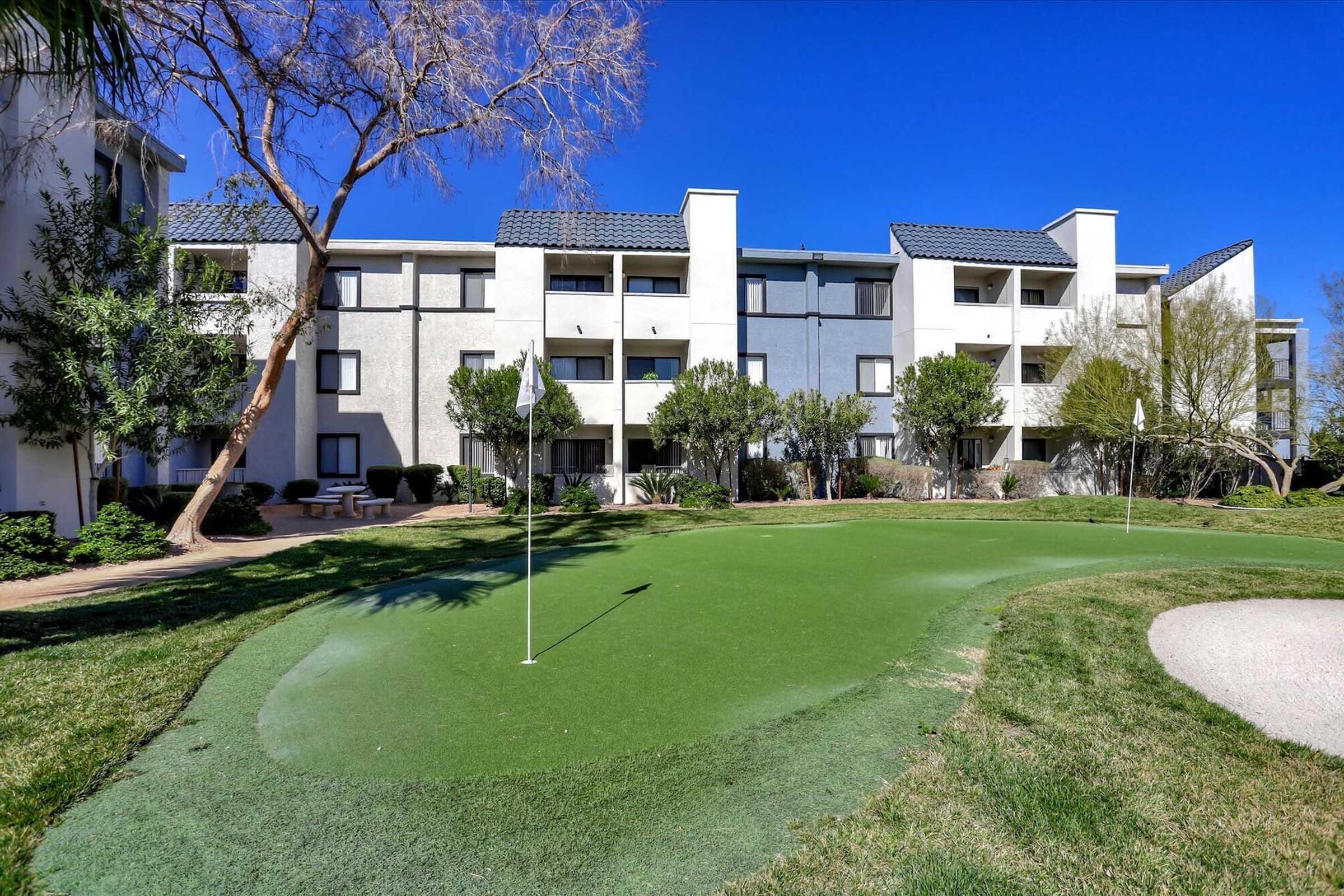 A well-maintained putting green with two flags in the foreground, surrounded by lush greenery. In the background, a modern residential building features balconies and large windows, under a clear blue sky.