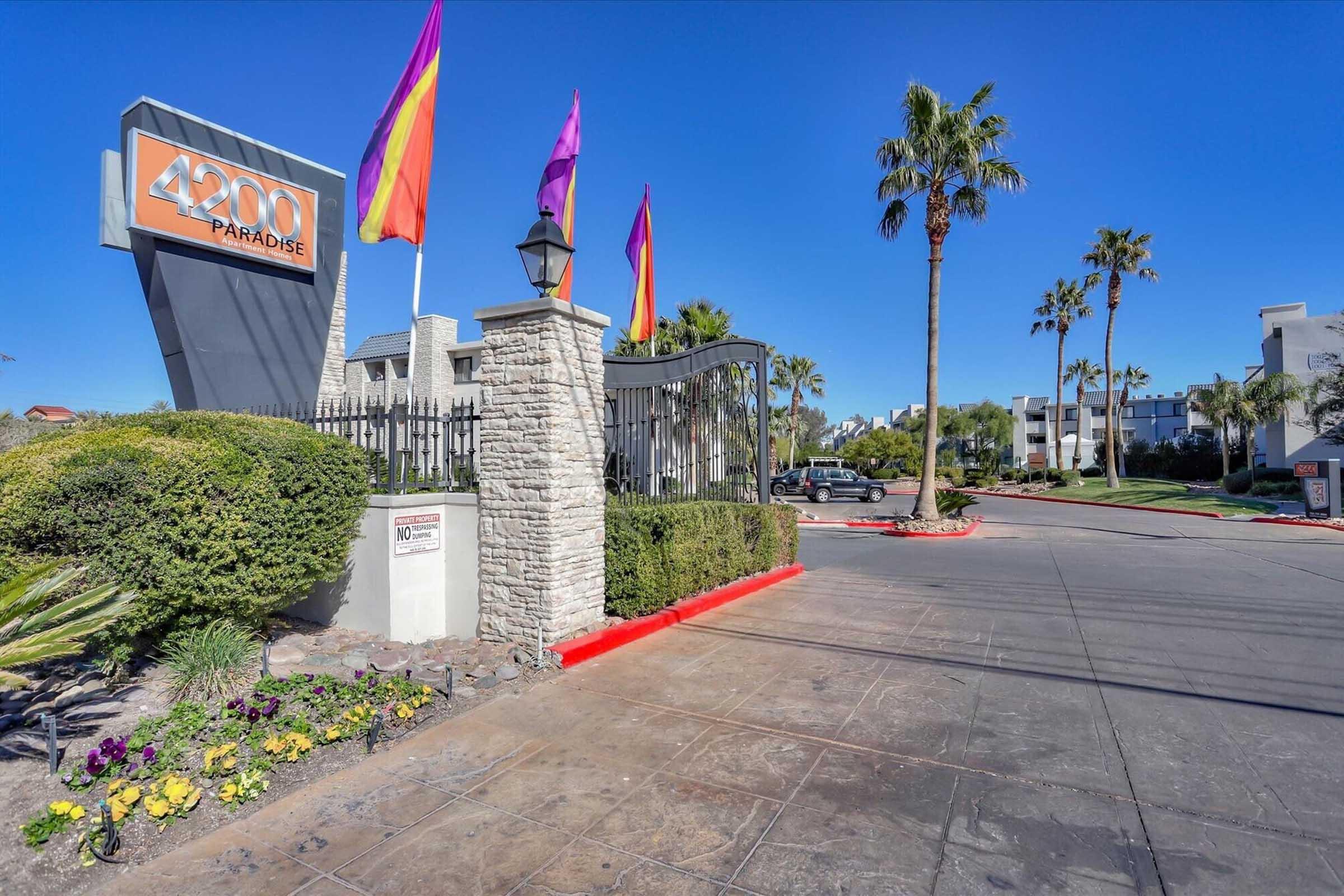 Sign for "4200 Paradise" at an entrance with colorful flags and palm trees. The pathway is lined with greenery and flowers, leading to an enclosed parking area. Clear blue sky in the background.
