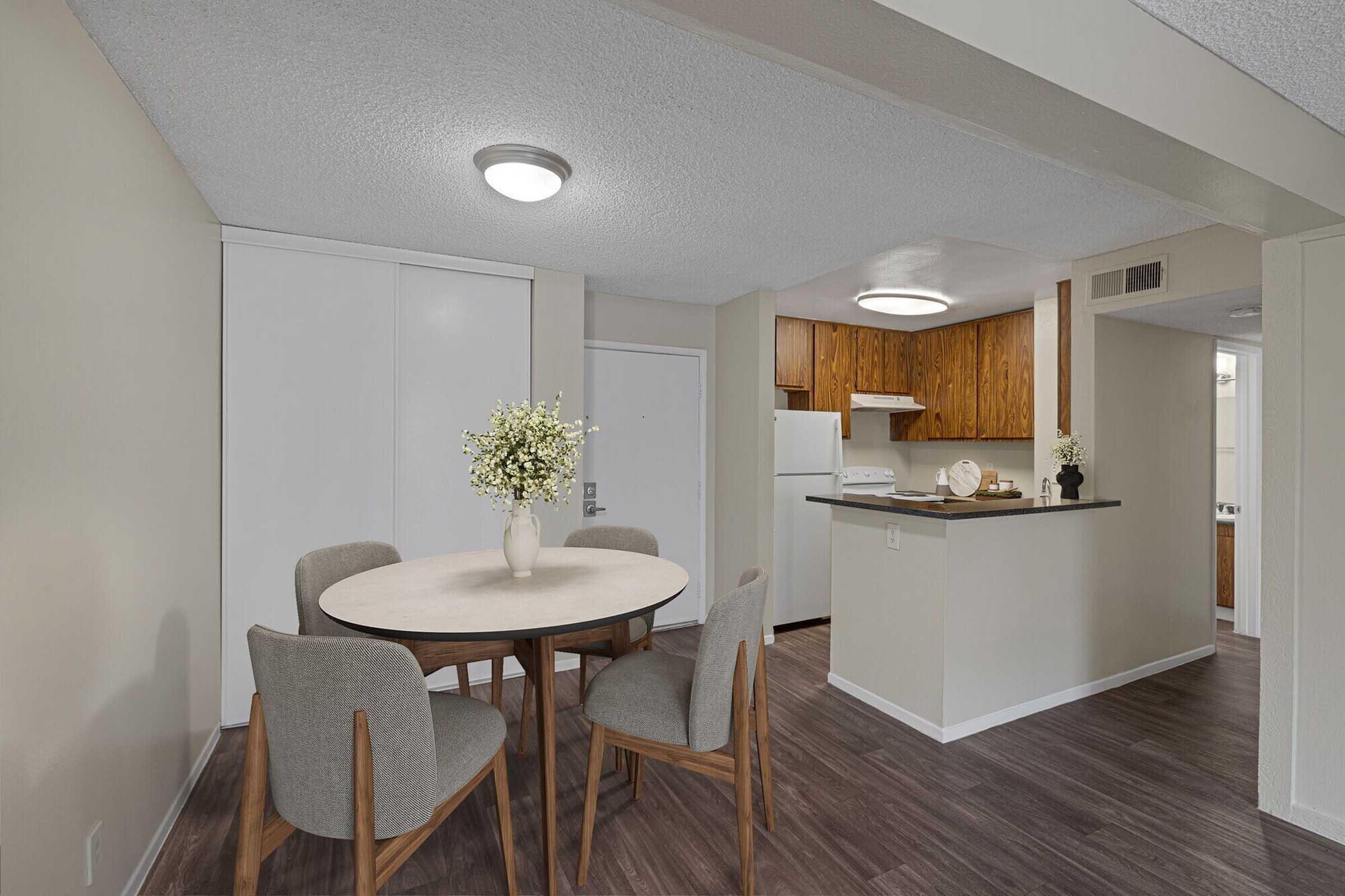 A modern dining area featuring a round table with four gray upholstered chairs, complemented by a small vase of flowers. In the background, a kitchenette with wooden cabinetry, a white refrigerator, and a neutral-colored wall creates a warm and inviting atmosphere. Natural light illuminates the space.