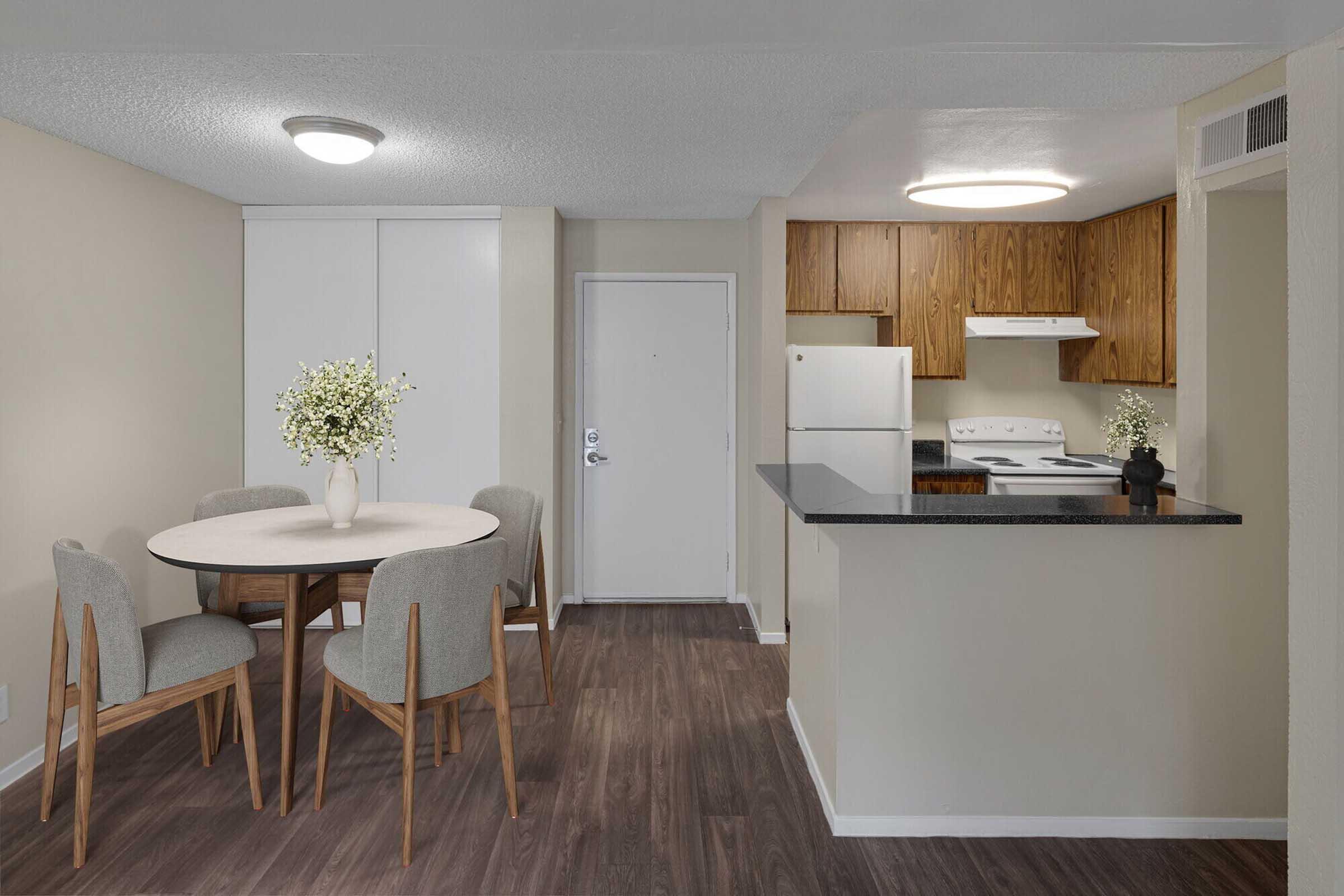 A bright, modern kitchen and dining area featuring a round wooden table with four upholstered chairs, a decorative vase with flowers, and a sleek countertop. The kitchen includes wood cabinets, a refrigerator, and a stove, with light-colored walls and a well-lit atmosphere.
