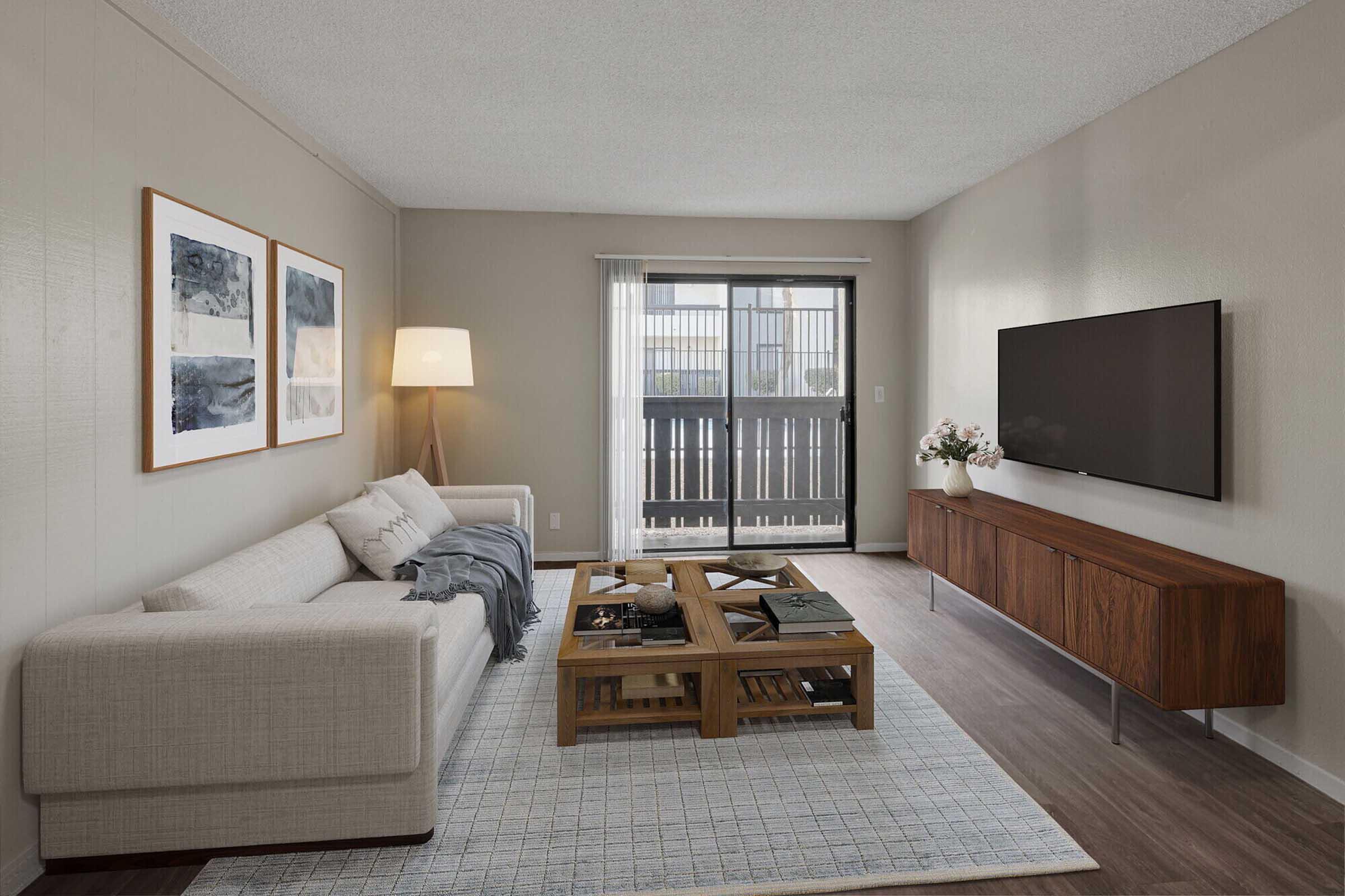 A modern living room featuring a light-colored sofa with a throw blanket, a wooden coffee table with decorative items, and a wooden TV stand. The walls are painted in a neutral color, and there are two framed artworks on the wall. A lamp stands beside the sofa, and large sliding glass doors lead to a balcony.