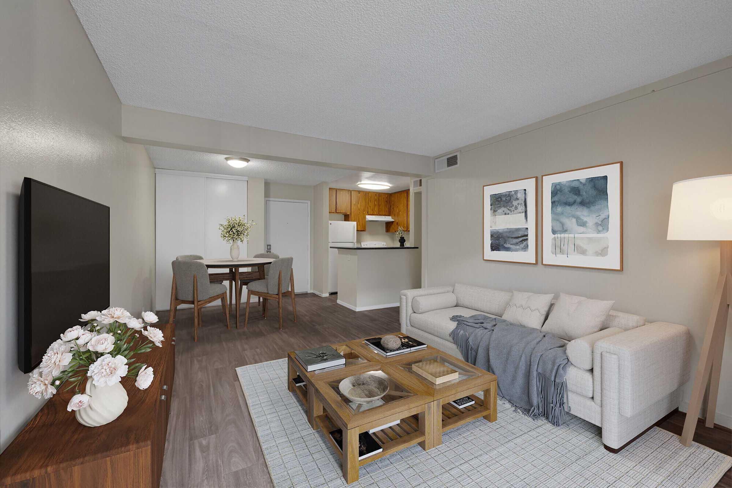 A modern living room featuring a light-colored sofa with a throw blanket, a wooden coffee table with decorative items, and a wall-mounted television. In the background, there is a dining area with a table and chairs, and a kitchen visible. The walls are painted in neutral tones, complemented by framed artwork.