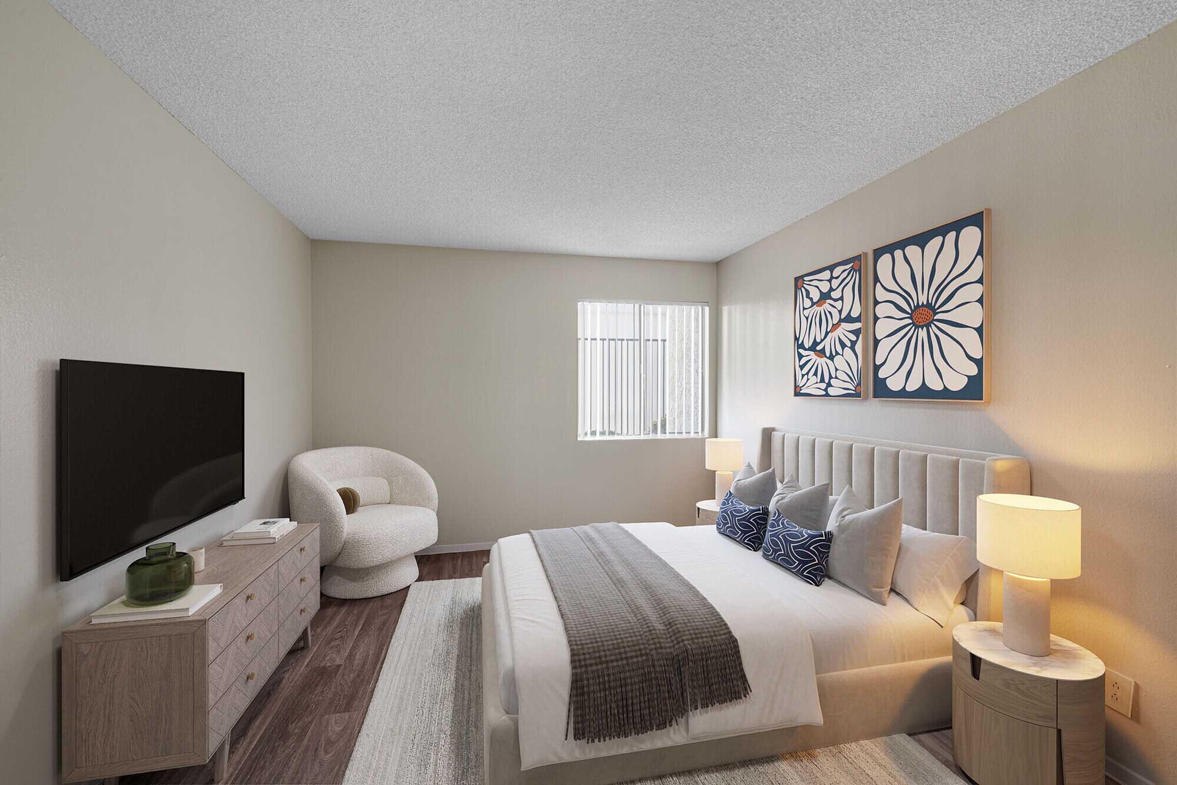 A modern bedroom featuring a light-colored wall, a large bed with decorative pillows, and two bedside lamps. There is a cozy armchair in the corner, a wooden dresser with decor items, a television on the wall opposite the bed, and two framed floral artworks. Natural light filters through a window with vertical blinds.