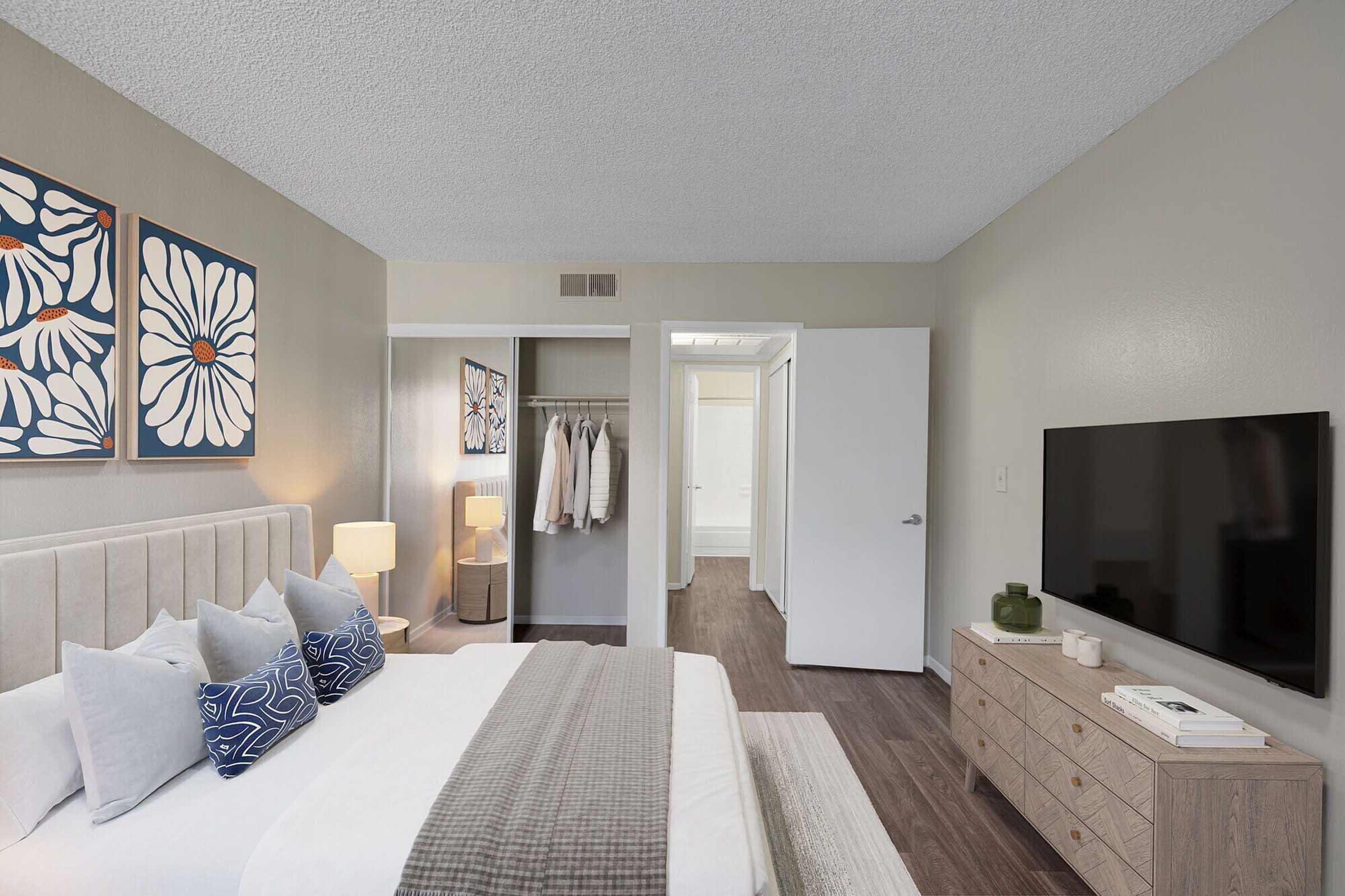 A modern bedroom featuring a king-sized bed with decorative pillows, a stylish bedside lamp, and floral artwork on the walls. A closet is seen to the left with hanging clothes, and a television is mounted across from the bed. The room has a neutral color palette and hardwood flooring. A doorway leads to an adjoining bathroom.