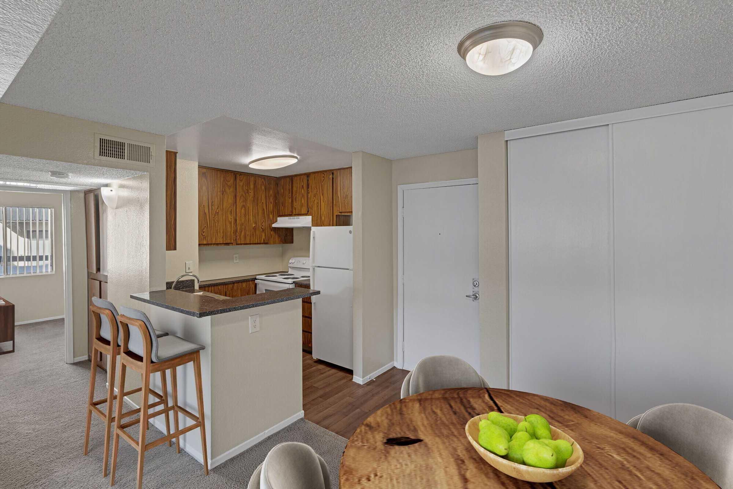 A modern apartment interior featuring a kitchen with wooden cabinets, a white refrigerator, and a dining area with a round wooden table and green apples. The space has light-colored walls, carpet flooring, and a large window providing natural light. Bar stools are positioned at the kitchen counter.