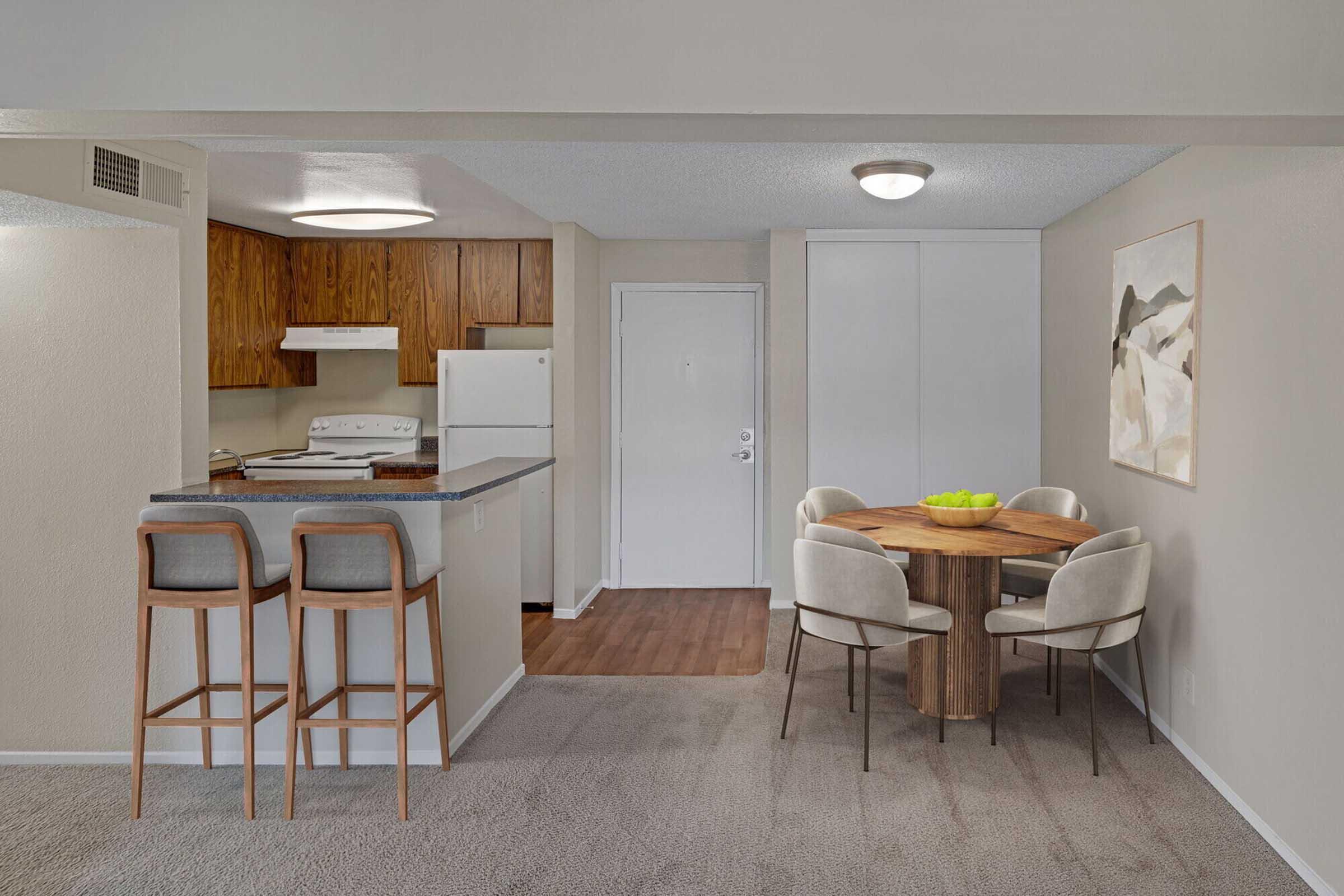 A modern kitchen and dining area featuring wooden cabinets, a white stove and refrigerator, and a round wooden dining table surrounded by four beige chairs. The space is well-lit with neutral-colored walls and a comfortable carpeted floor. A bowl of fruit is positioned on the table.
