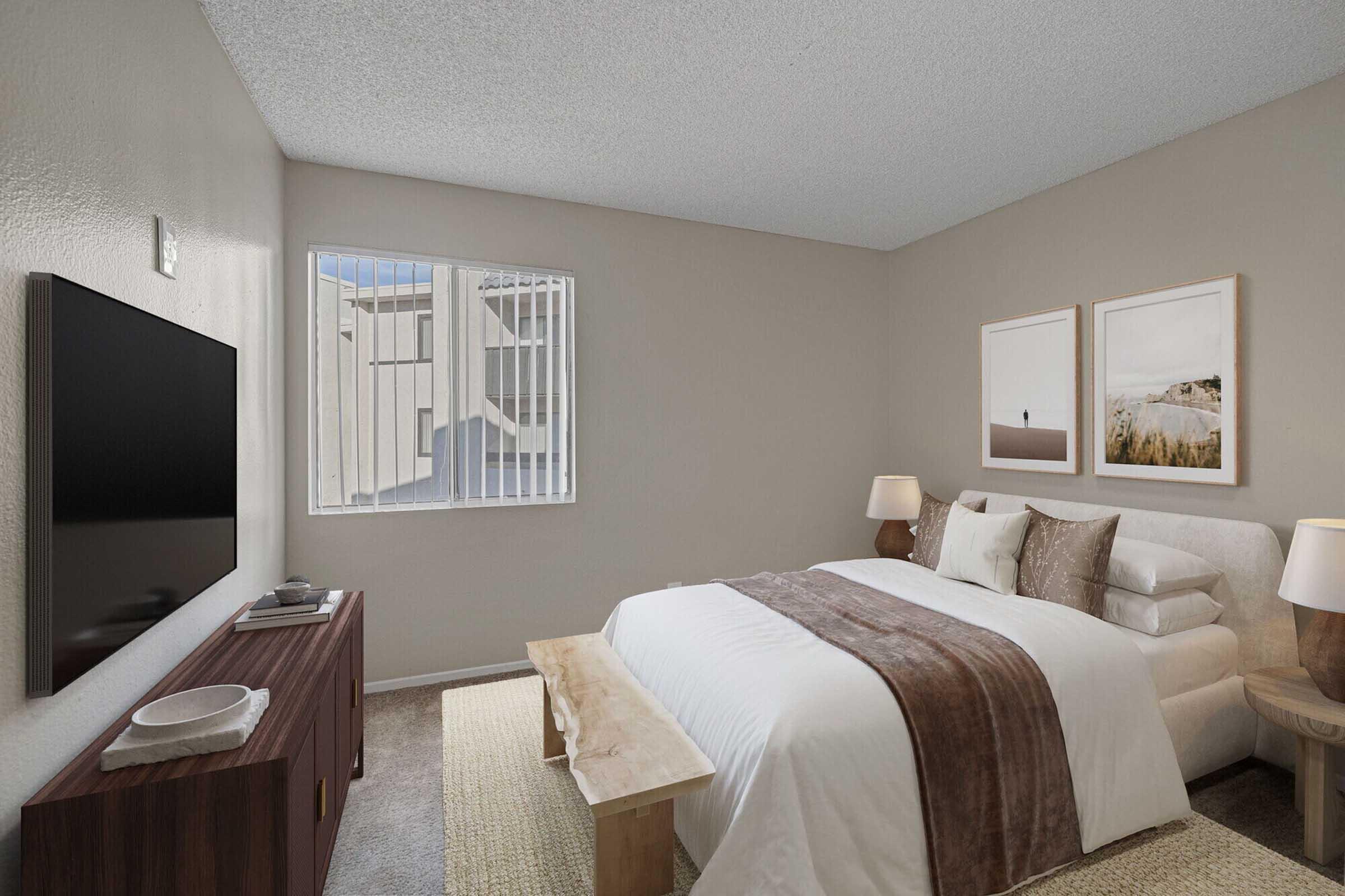 Cozy bedroom featuring a large bed with a light-colored bedding ensemble, a wooden bench at the foot of the bed, and modern bedside lamps. A TV is mounted on the wall opposite the bed, with a wooden dresser below it. Natural light comes through a window with vertical blinds, and neutral-toned walls enhance the serene atmosphere.