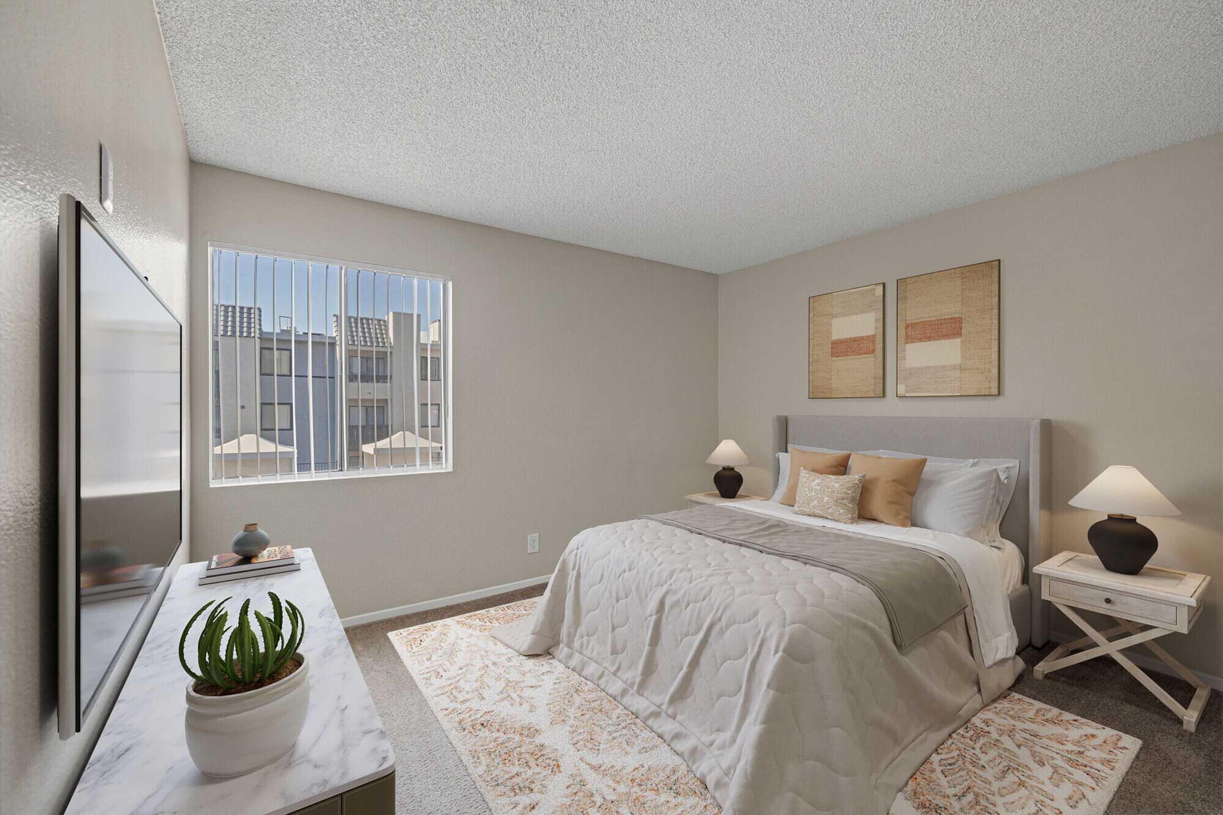 A cozy bedroom featuring a queen-sized bed with white bedding and decorative pillows. There are two framed artworks on the wall above the bed. A television is mounted on the opposite wall, alongside a white dresser with a small potted plant. Natural light filters through a window with vertical blinds.