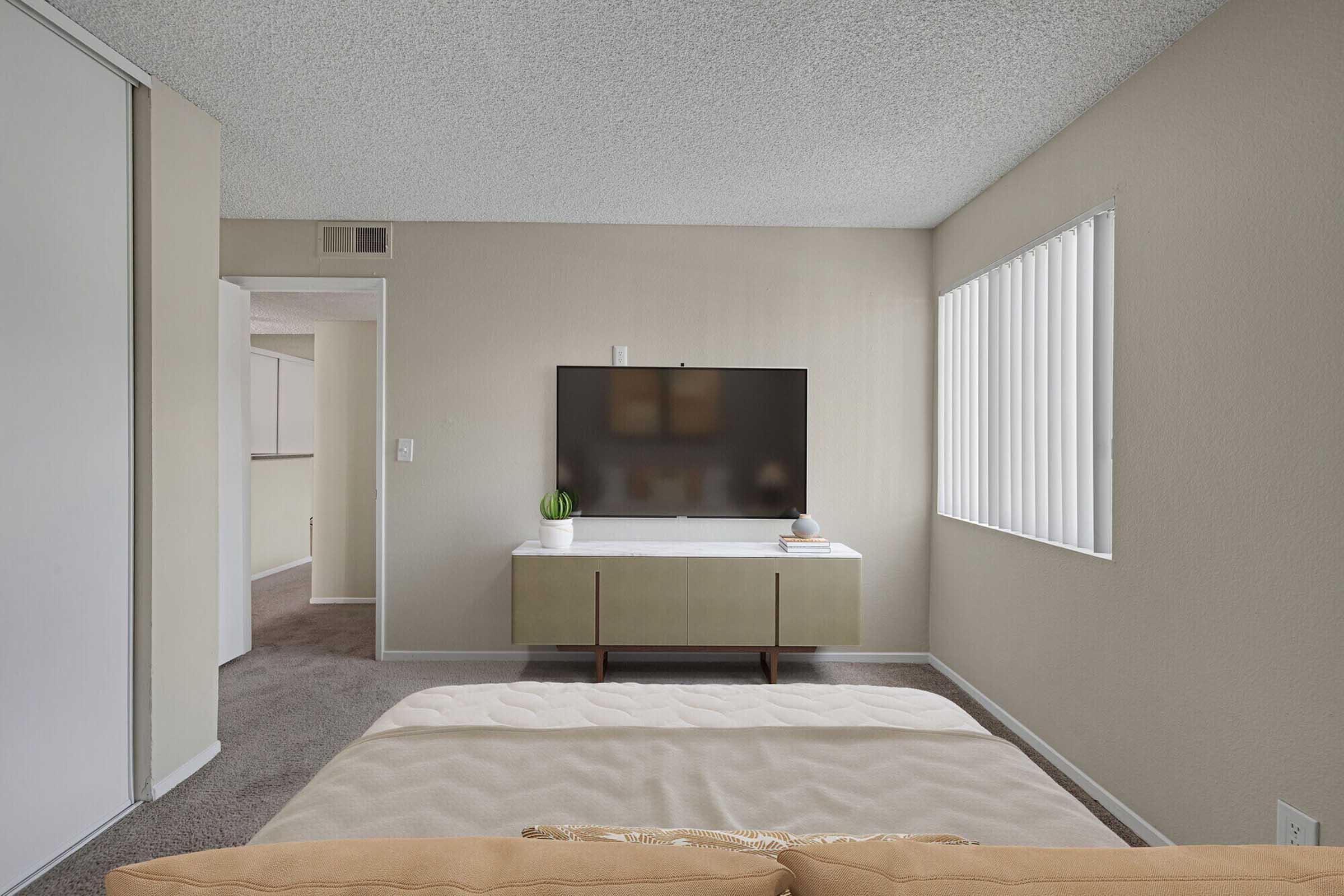 A cozy bedroom with a light-colored bedspread in the foreground. A modern TV mounted on the wall opposite the bed, with a stylish media console underneath. Neutral-toned walls and carpet create a warm atmosphere, and vertical blinds allow soft light to filter in. A glimpse of an adjoining room is visible on the left.