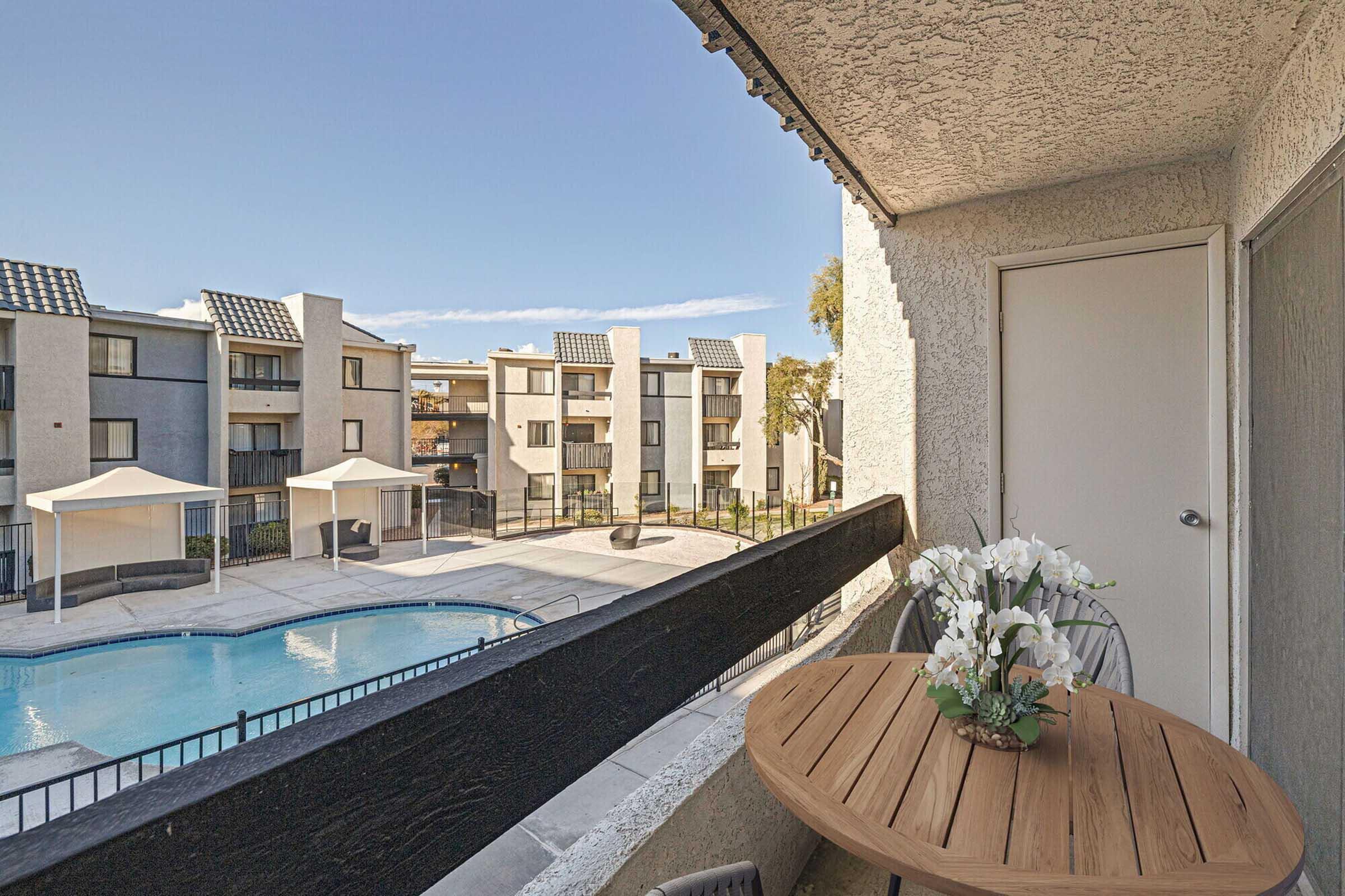 View from a balcony overlooking a swimming pool and courtyard. The scene features a round wooden table with a vase of flowers, a few lounge chairs, and several apartment buildings in the background, each with a patio and solar panels on the roofs, under a clear blue sky.