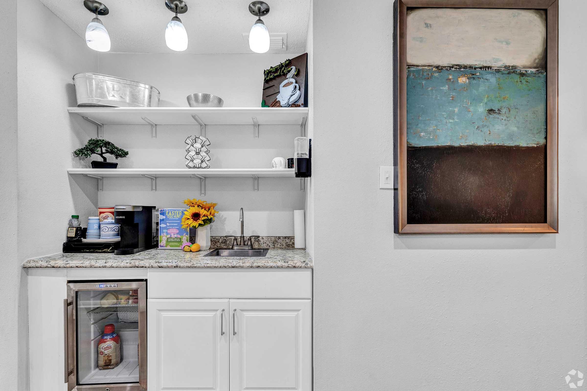 A kitchen nook featuring a small counter with a sink and various items, including a coffee maker, a decorated wall shelf, and a mini fridge. A painting is hung on the wall above, showcasing abstract colors of blue, brown, and beige, while a vase with sunflowers adds a touch of warmth to the space.