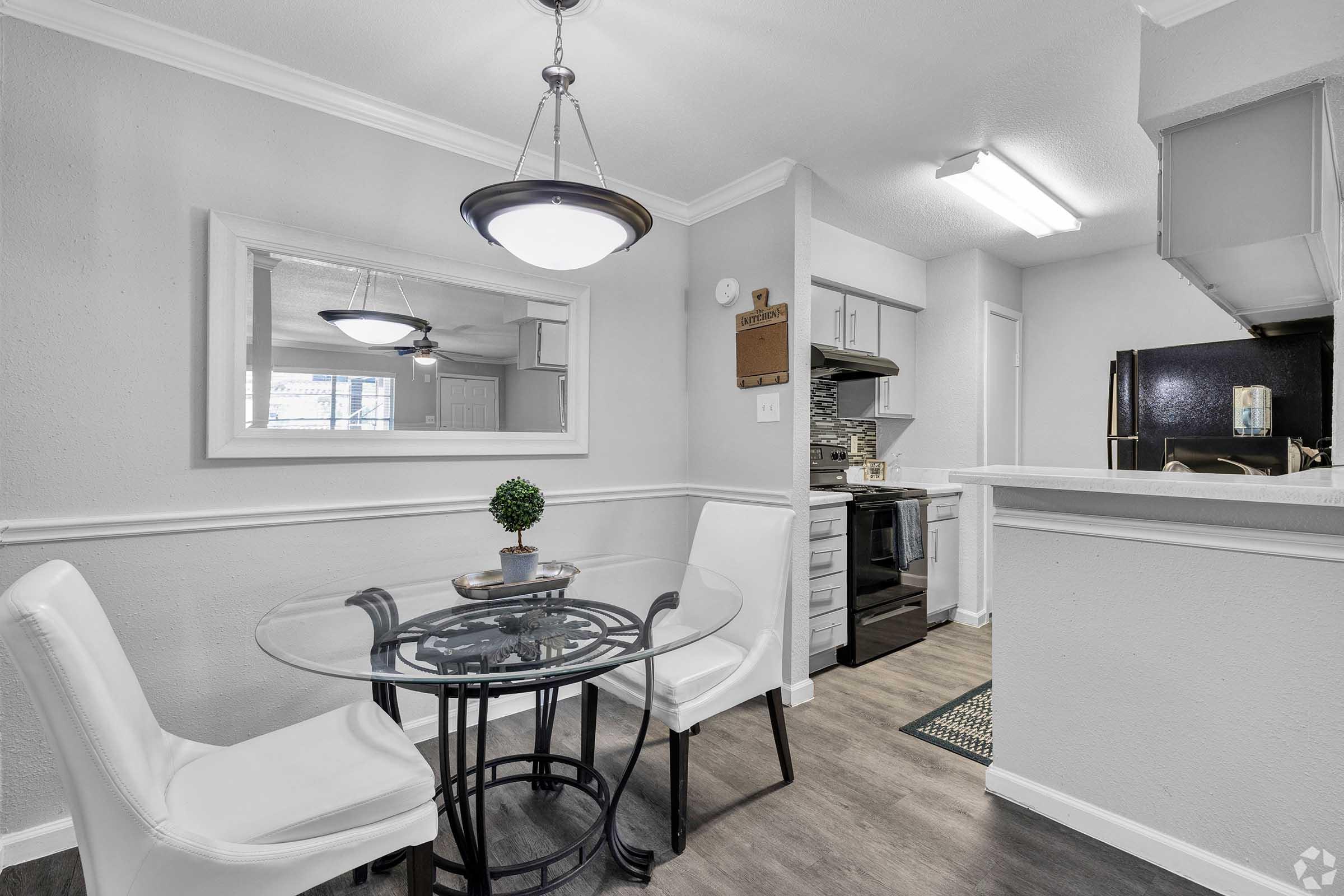 A modern kitchen and dining area featuring a white round table with two white chairs, a decorative plant on the table, a large mirror on the wall, and a well-equipped kitchen visible in the background with dark cabinets and appliances.
