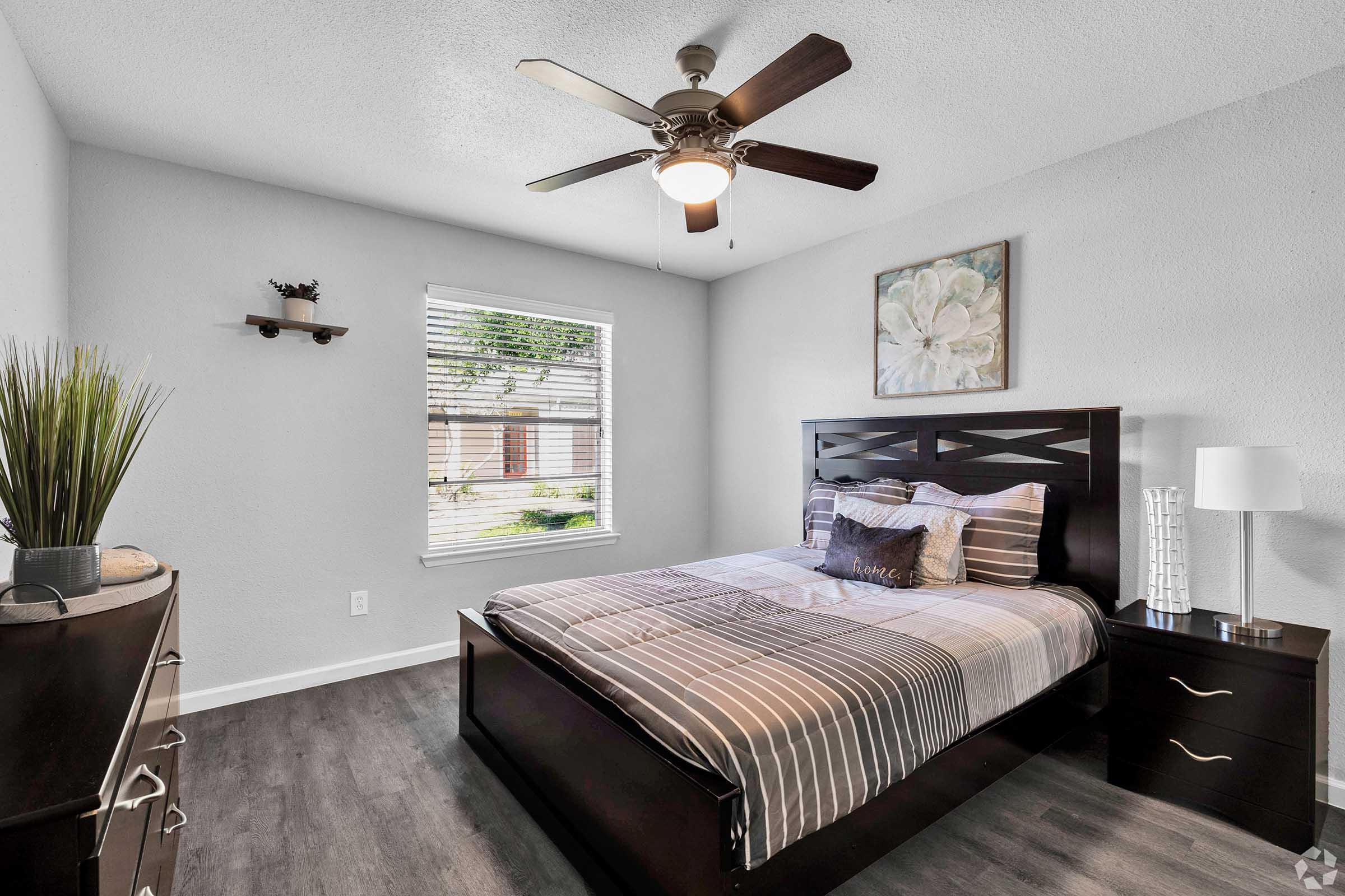 A modern bedroom featuring a queen-sized bed with striped bedding, a bedside table with a lamp, and a dresser. A ceiling fan is installed above, and a window allows natural light to enter, complemented by decorative plants and a wall-mounted shelf with small decor items.