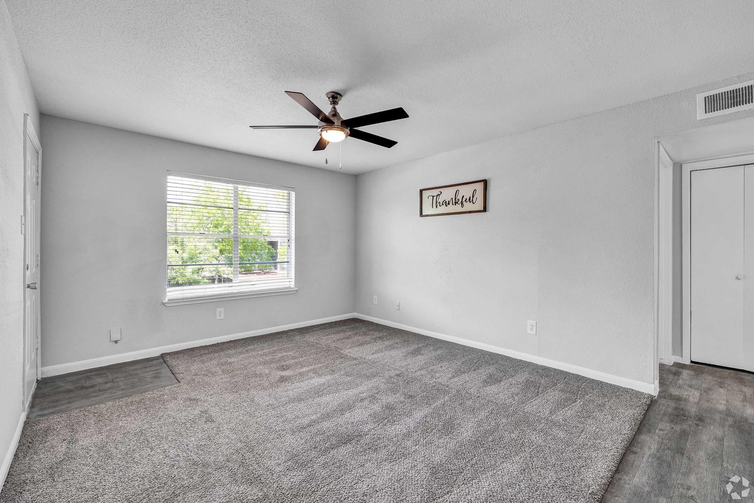 A well-lit room with a ceiling fan and a large window. The walls are painted light gray, and the flooring features gray carpet. A decorative sign that says "Thoroughbred" is mounted on one wall, and there's a white closet door visible in the corner.