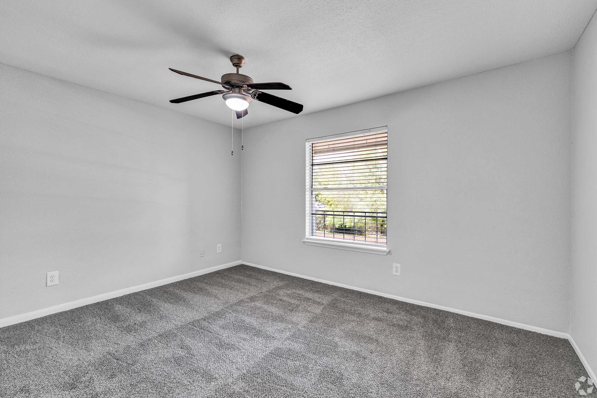 A spacious, empty room with gray carpet and light gray walls. It features a ceiling fan with a light fixture and a window with blinds, offering natural light. The room has a clean and modern feel, ideal for personalization.