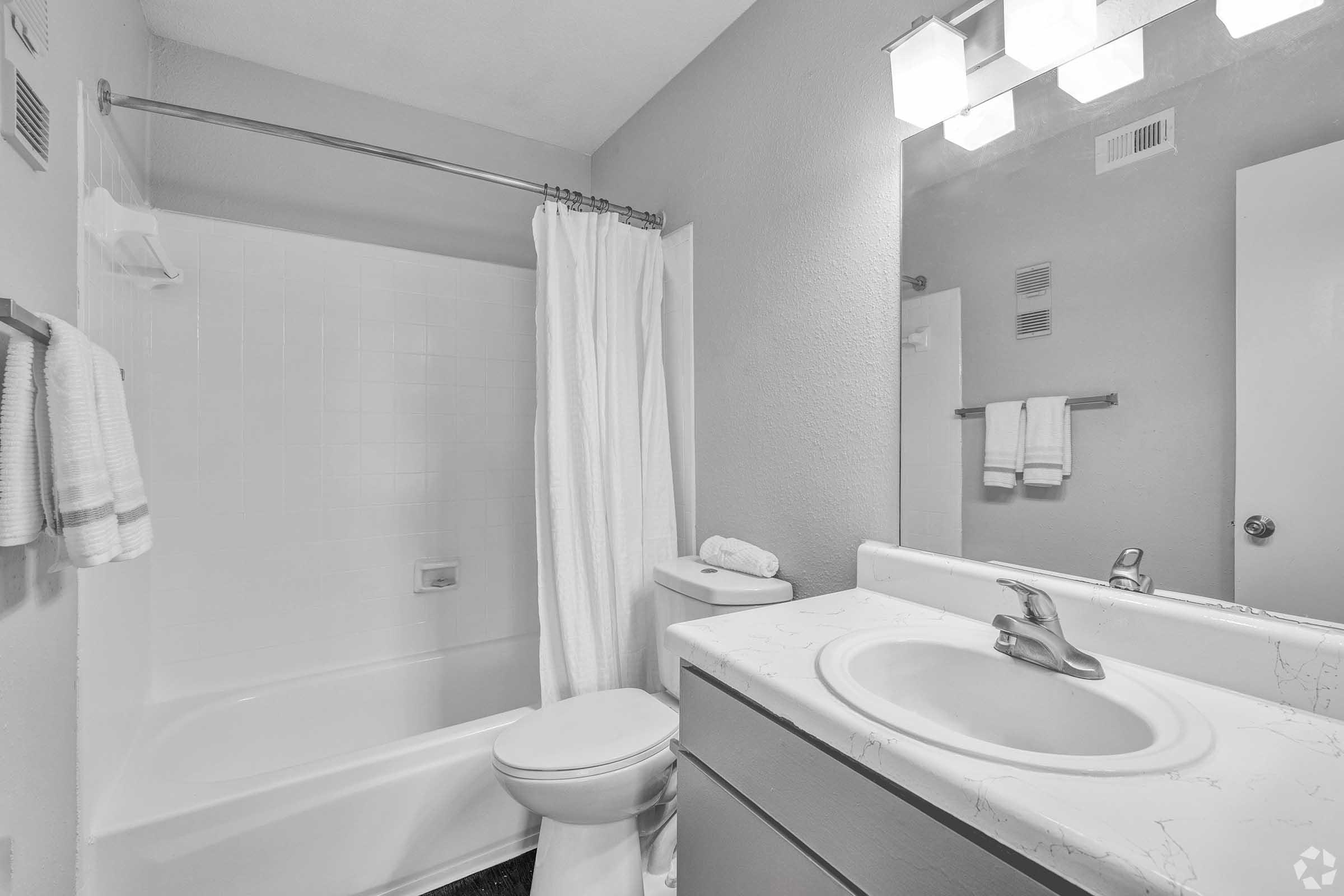 A clean, minimalist bathroom featuring a bathtub with a shower curtain, a toilet, and a sink with a marble countertop. Soft lighting from a wall-mounted mirror fixture complements the neutral-colored walls. Two towels hang neatly on a bar beside the shower.