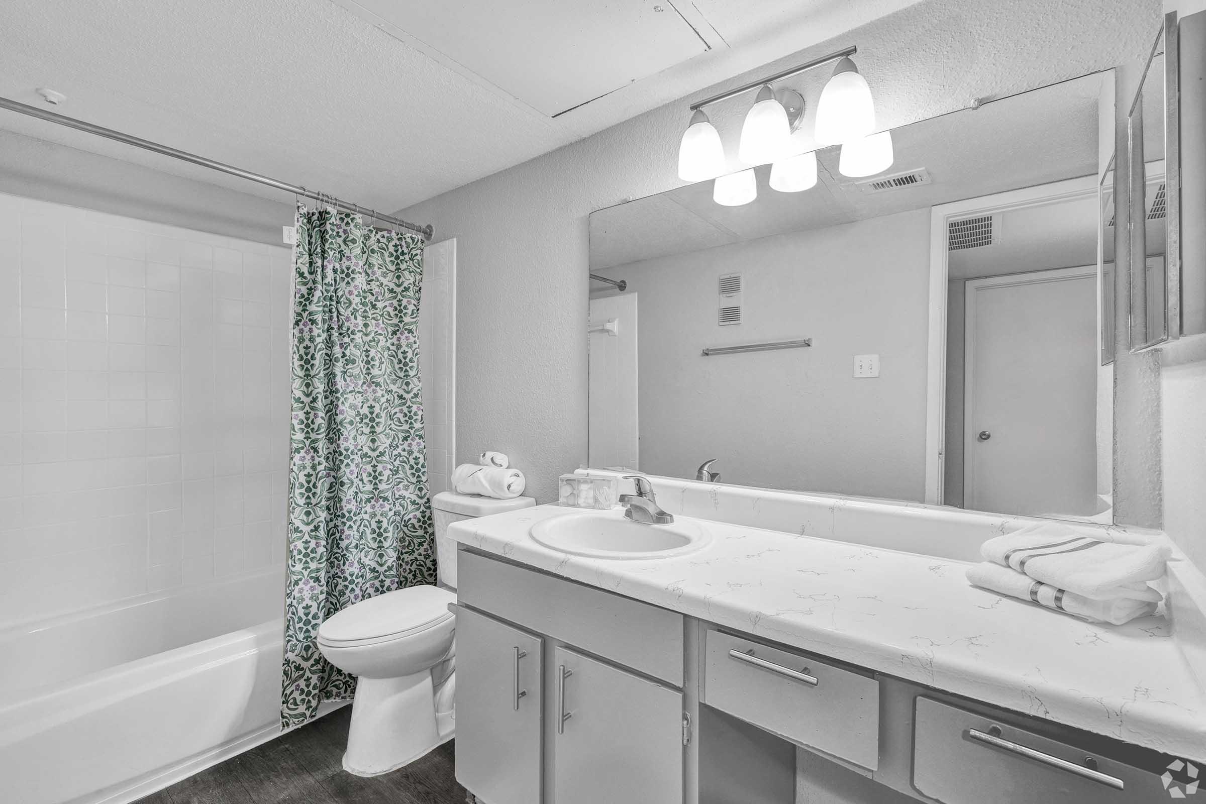 A simple bathroom featuring a white bathtub with a shower curtain displaying a floral pattern, a white sink countertop with toiletries, a large mirror above the sink, and two light fixtures. There are neatly stacked towels on the countertop and a minimalist decor style.