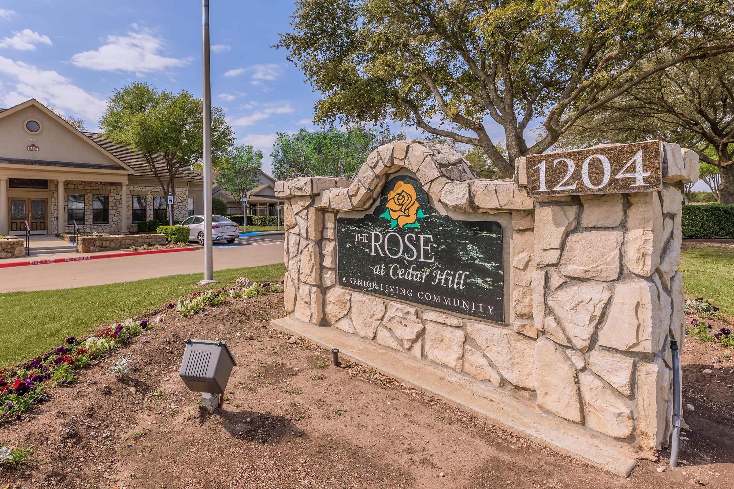 Sign for "The Rose at Cedar Hill," a senior living community. The stone monument features decorative landscaping with flowers, indicating the entrance to the facility. A clear blue sky is visible in the background, along with nearby buildings and parked cars.