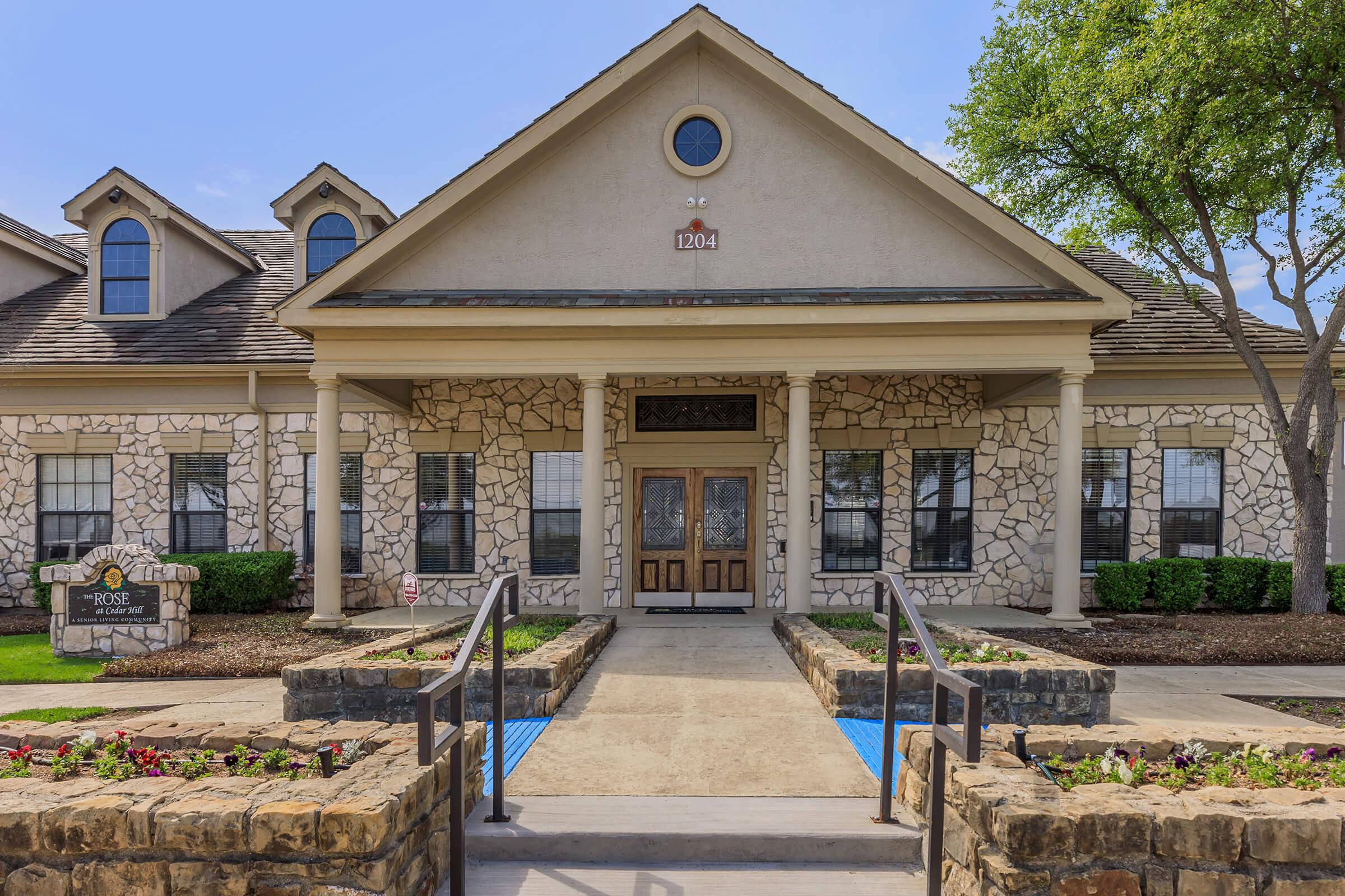 Front view of a two-story building with a stone facade, featuring a central entrance with double wooden doors. The pathway leading to the entrance is lined with flowers and has two sets of steps. Large windows flank the entrance, and there are green shrubs and trees around the property. A clear blue sky is visible overhead.