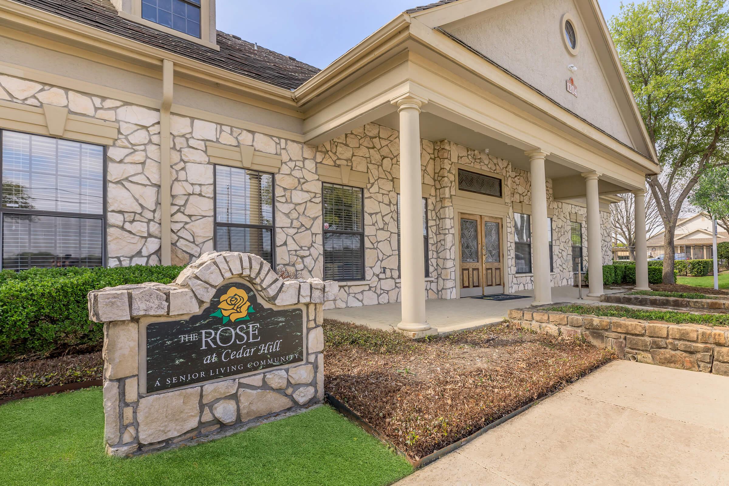 Exterior view of a senior living community named "The Rose at Cedar Hill," featuring a stone facade and well-maintained landscaping. A prominent sign at the entrance displays the community's name, highlighting its welcoming atmosphere.