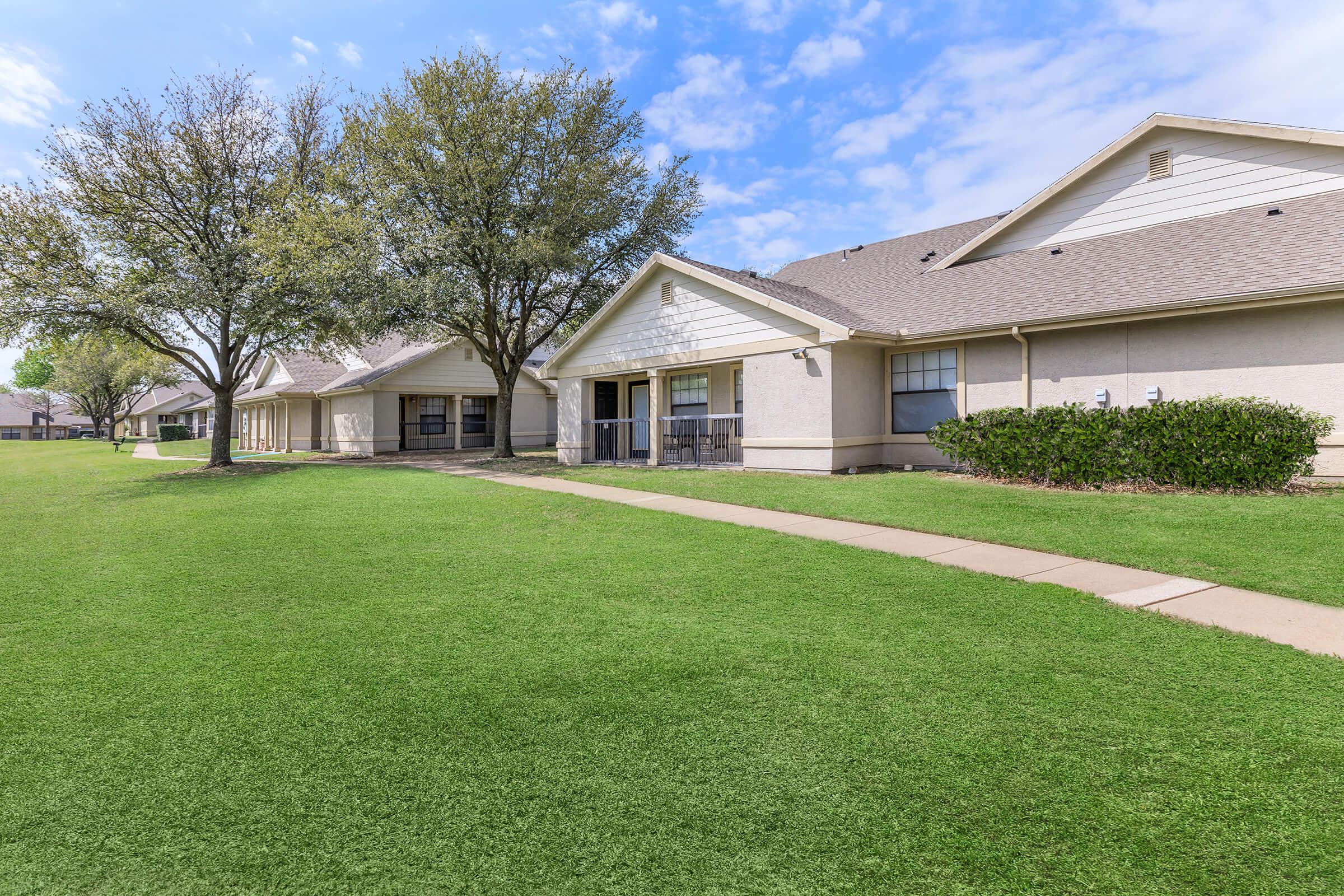 A grassy area with well-maintained sidewalks leading to single-story residential buildings. Trees line the path, and the sky is partly cloudy. The scene conveys a peaceful suburban atmosphere.