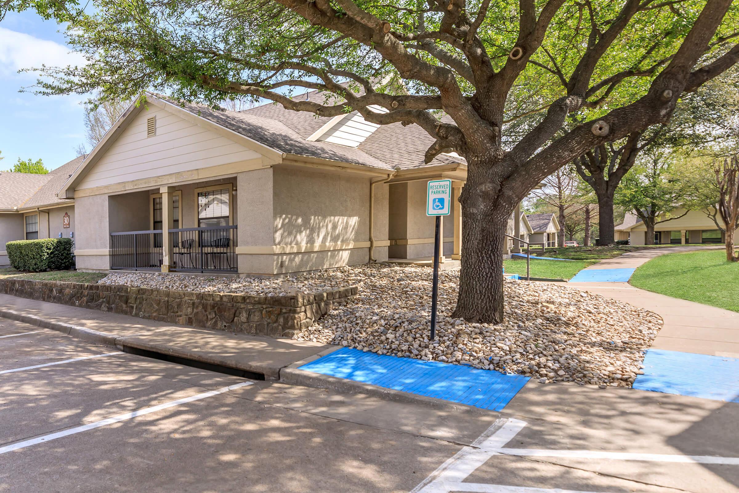 A residential building with a gray exterior and a porch, surrounded by landscaped rocks and trees. There is a designated parking space for people with disabilities, marked by a sign. A blue pathway leads through the grass, enhancing accessibility.
