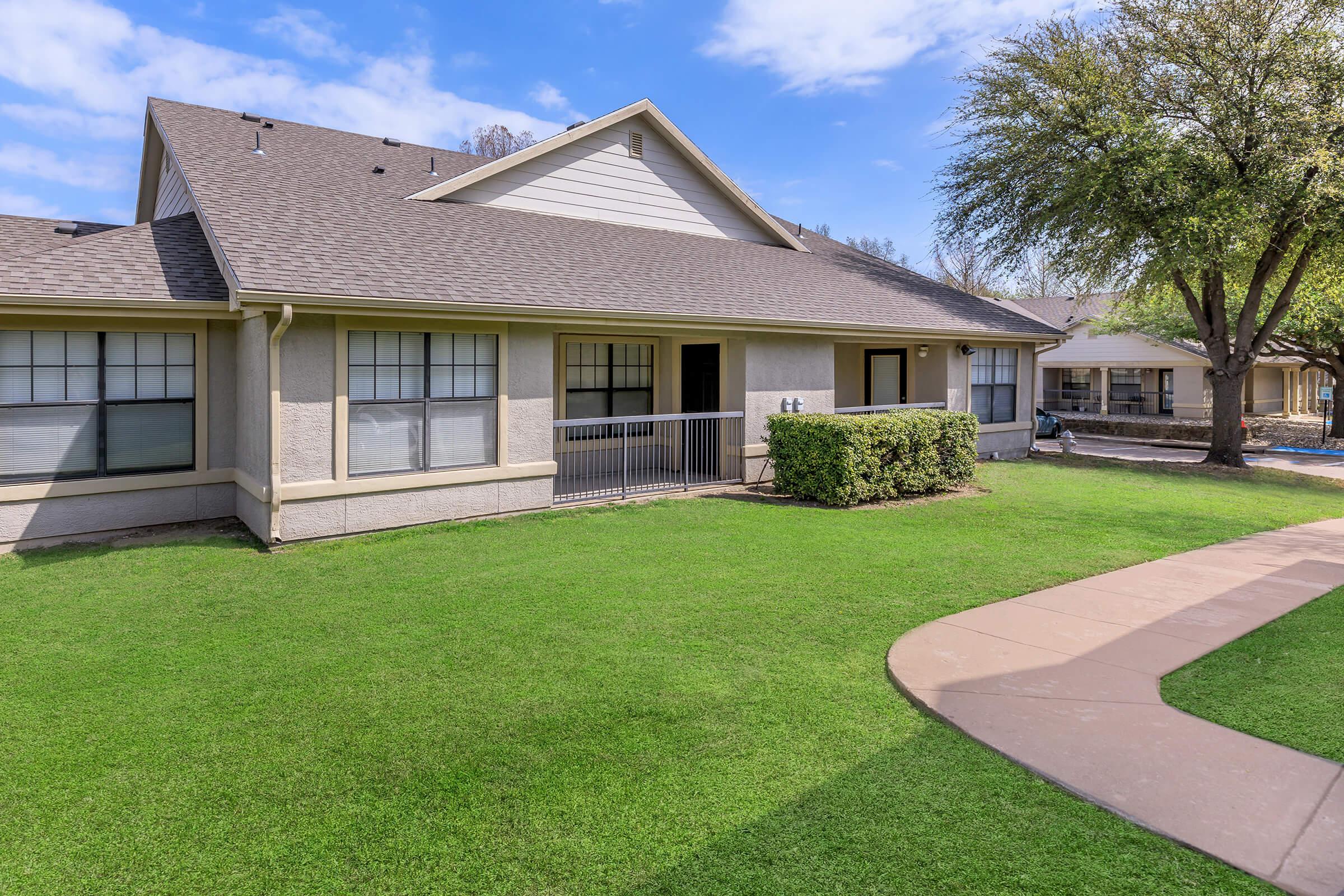 A residential building featuring a light-colored exterior, multiple windows, and a small porch area. The surrounding yard is well-maintained with green grass and a pathway leading to the right. Trees and additional outdoor space can be seen in the background under a partly cloudy sky.