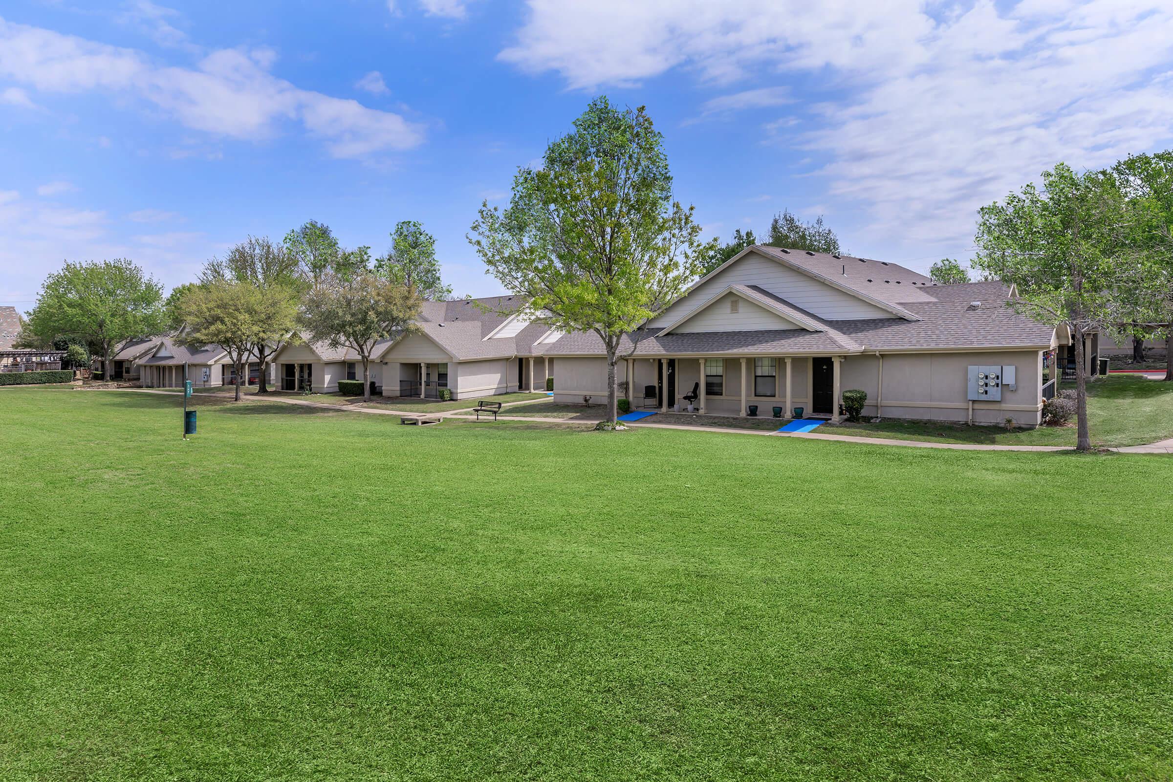 A well-maintained grassy area with several single-story residential buildings. Each building has a porch and is surrounded by trees and landscaping. The sky is clear with a few clouds, indicating a bright day. The scene conveys a peaceful and inviting neighborhood atmosphere.