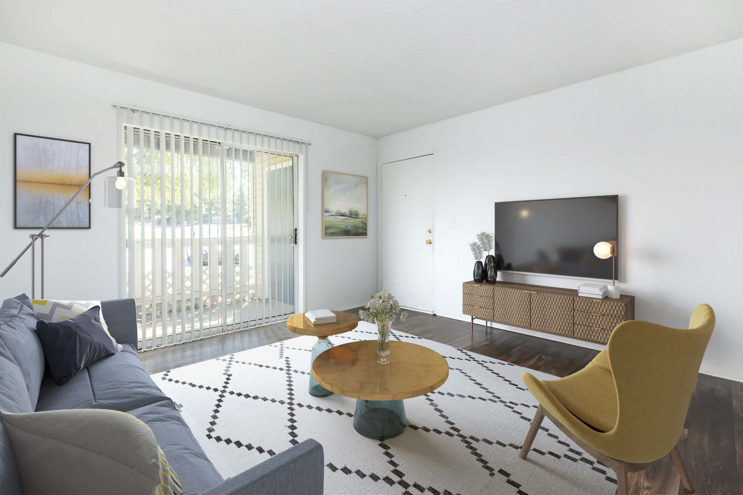 A bright, modern living room featuring a blue sofa with colorful cushions, a round wooden coffee table, and a yellow accent chair. There is a large flat-screen TV on a wooden console, and the space is decorated with light-colored art pieces and a patterned rug. Large sliding glass doors provide natural light.