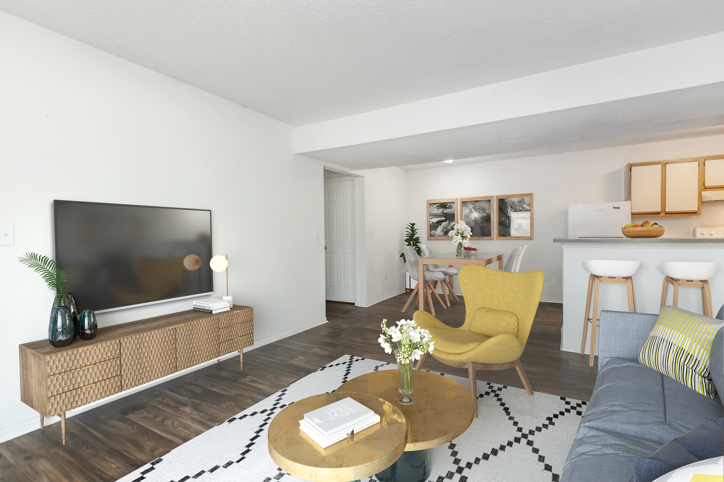 Bright and modern living area featuring a gray sofa and a yellow accent chair. A wooden coffee table sits in the center, with a vase of flowers. A TV is mounted on the wall above a stylish media console. In the background, a dining area with a circular table and bar stools is visible, enhancing the open layout.