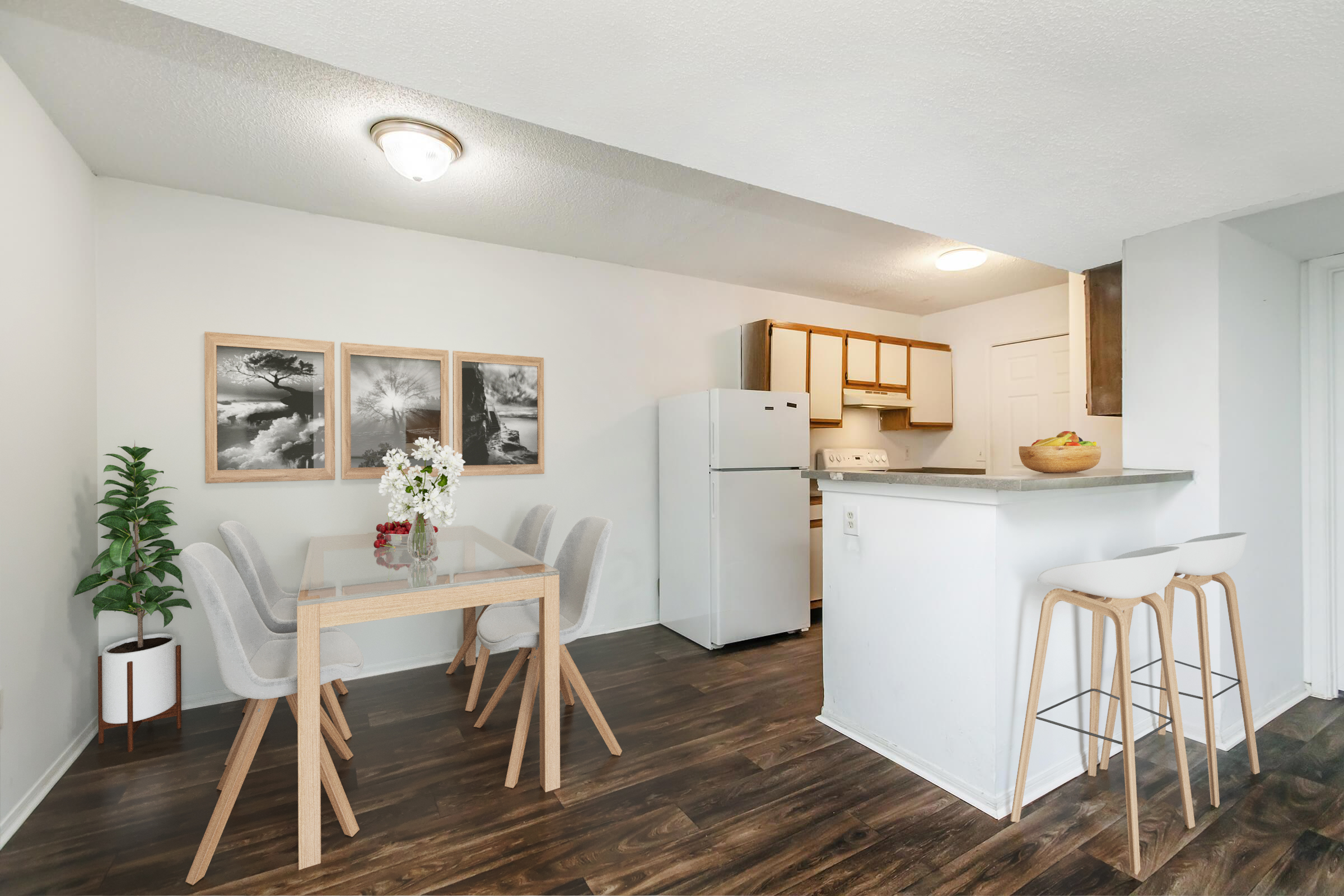 A modern kitchen and dining area featuring a dining table with four chairs, a refrigerator, and cabinets. Two framed pictures hang on the wall, and a potted plant adds greenery. Bar stools are positioned near a countertop, and the floor has a wood-like finish, creating a welcoming atmosphere.
