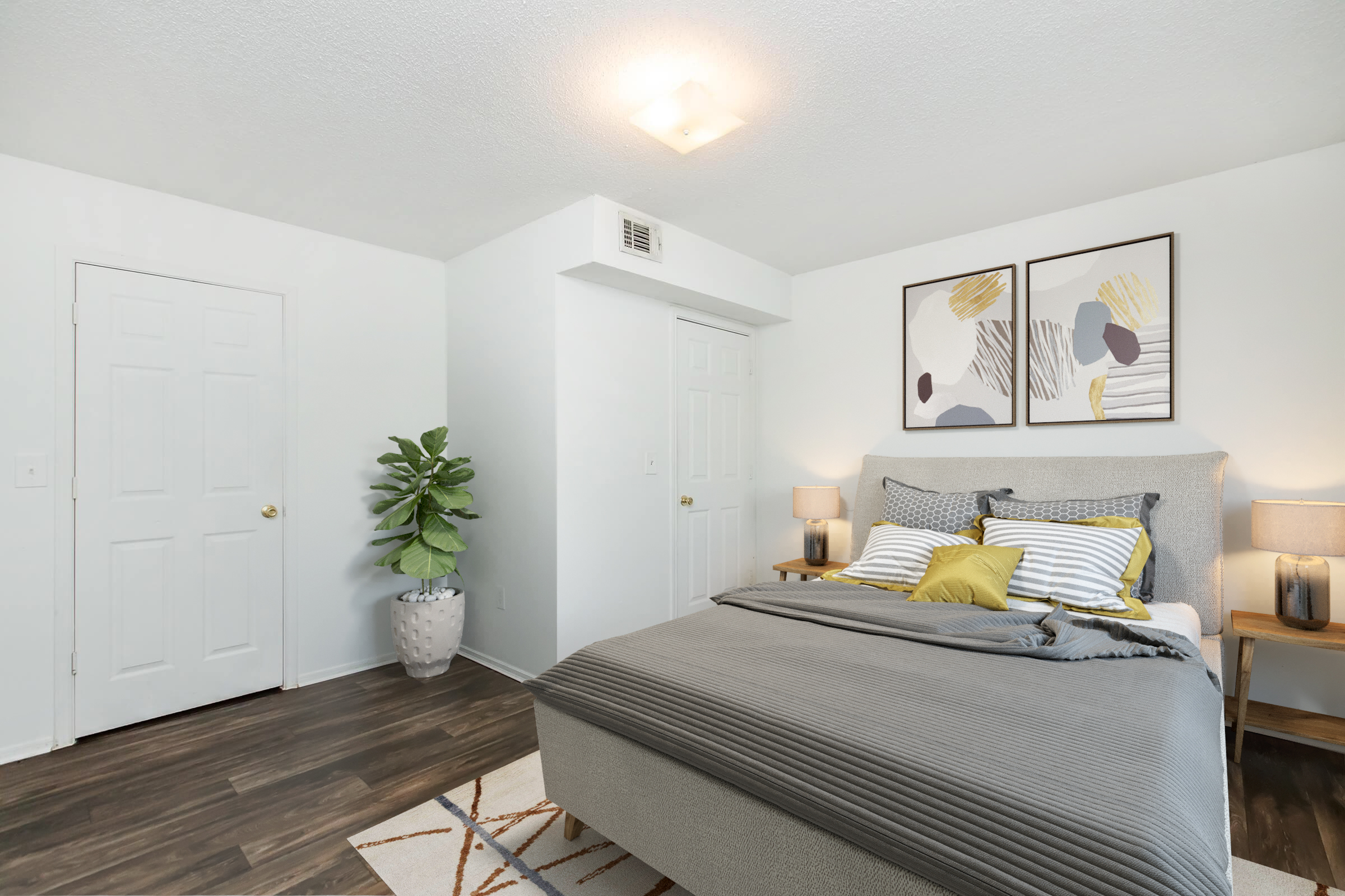 Bright and airy bedroom featuring a gray upholstered bed with yellow and white accent pillows. Two bedside tables with lamps flank the bed. A large wall art adorns the wall above the bed, and a potted plant adds greenery. A closed door is visible on the left, and another door leads to a closet on the right.