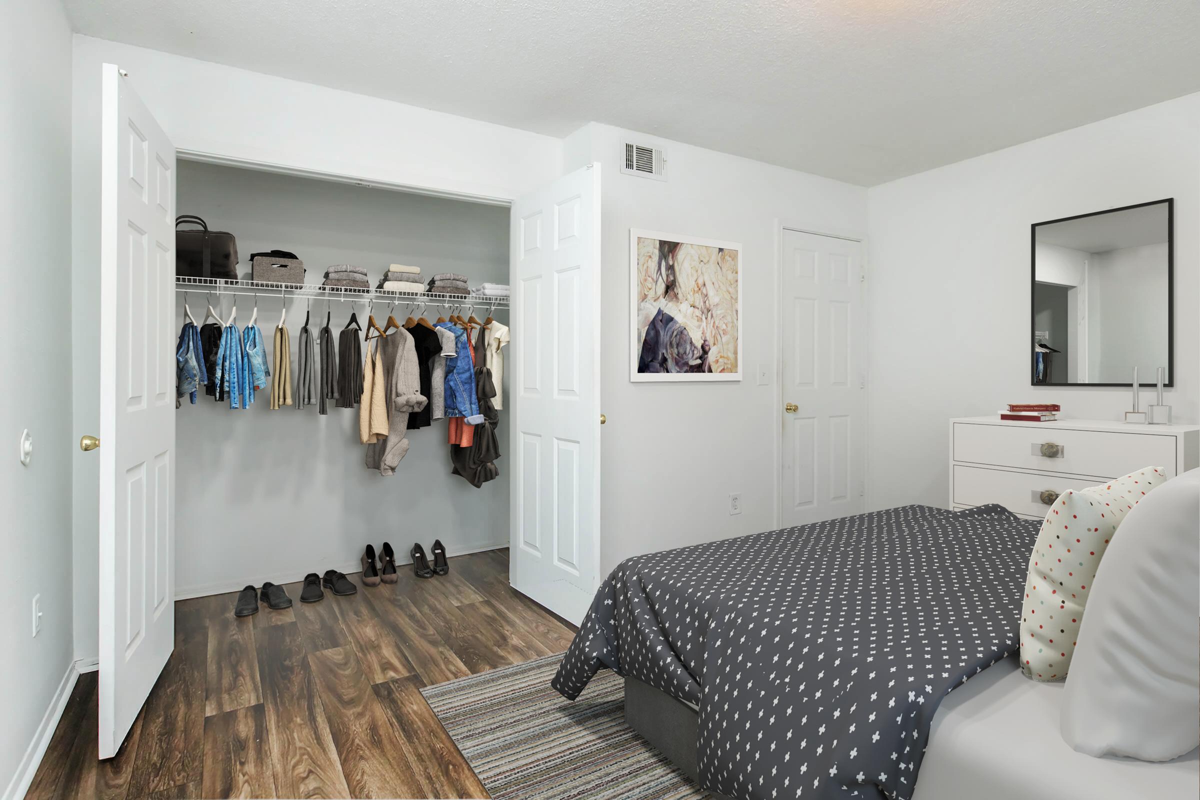 A modern bedroom featuring a neatly made bed with a patterned duvet cover, a small area rug, and a white dresser with a mirror. An open closet displays neatly hung clothing and shoes on the floor, emphasizing a tidy, minimalist aesthetic with light-colored walls and wooden flooring.