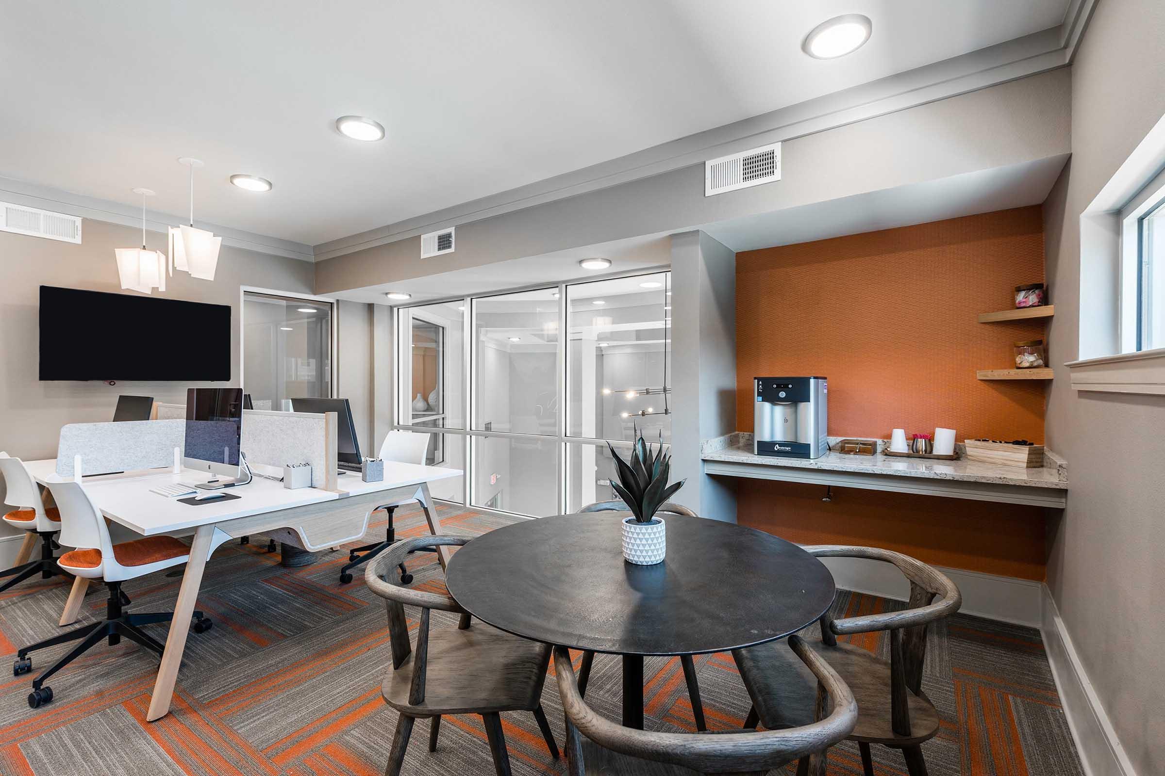 A modern, well-lit office space featuring a sleek work desk with multiple workstations, a round dining table with chairs, and a coffee station.