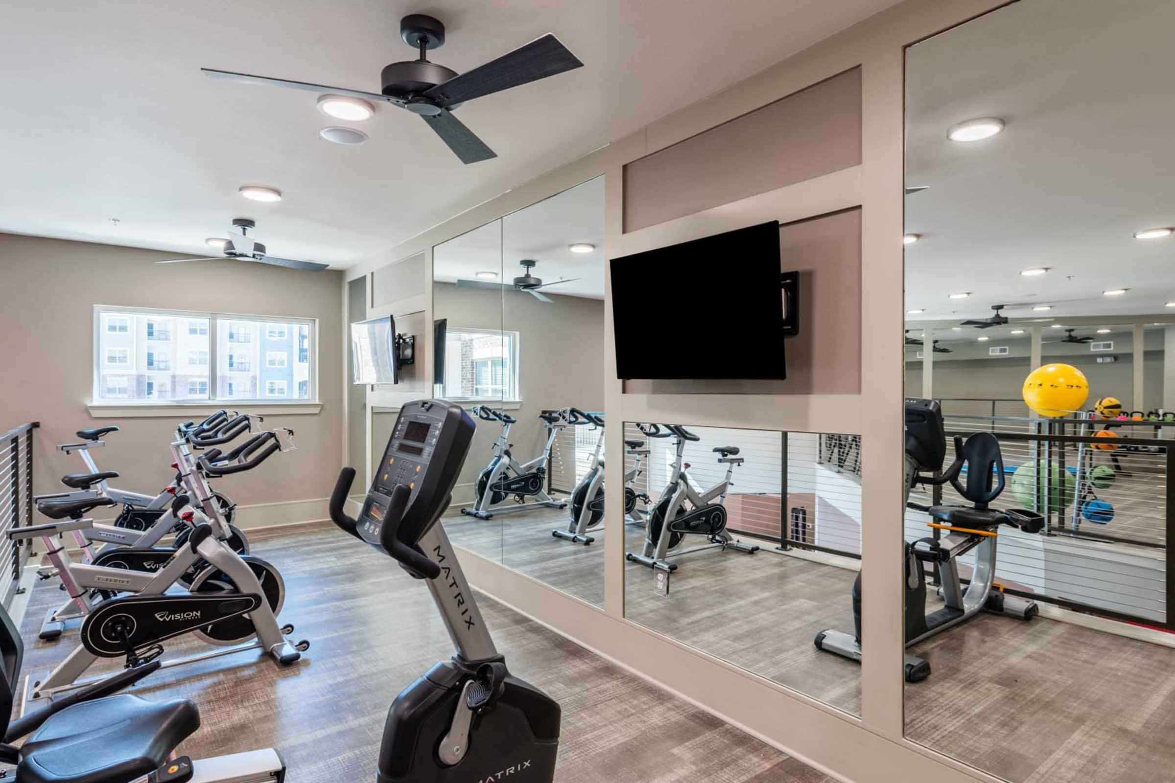 A modern fitness room featuring exercise equipment including stationary bikes. The space has large mirrors, ceiling