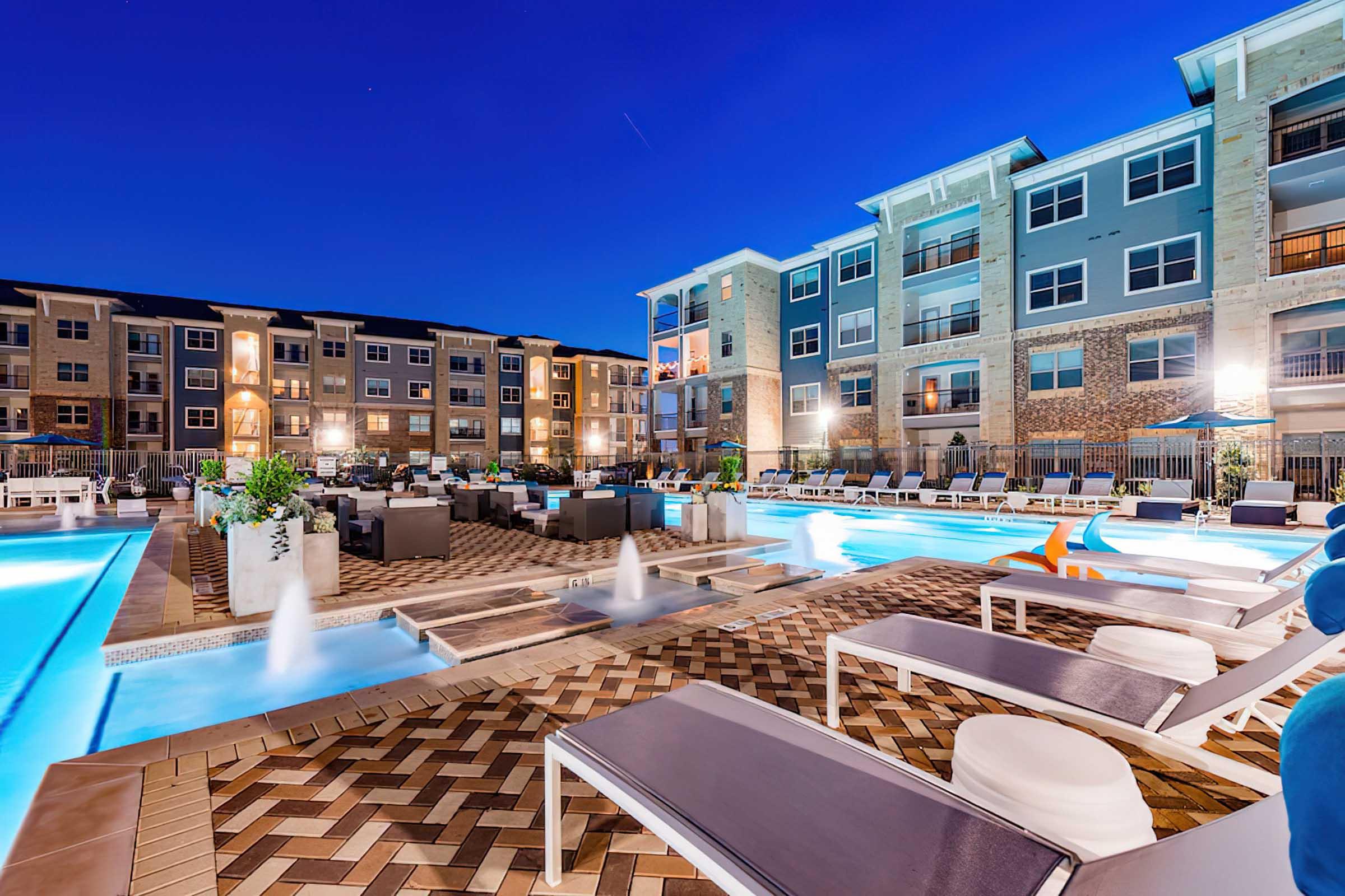 A modern apartment complex at night with a well-lit pool area. The pool features fountains and is surrounded by lounge chairs.
